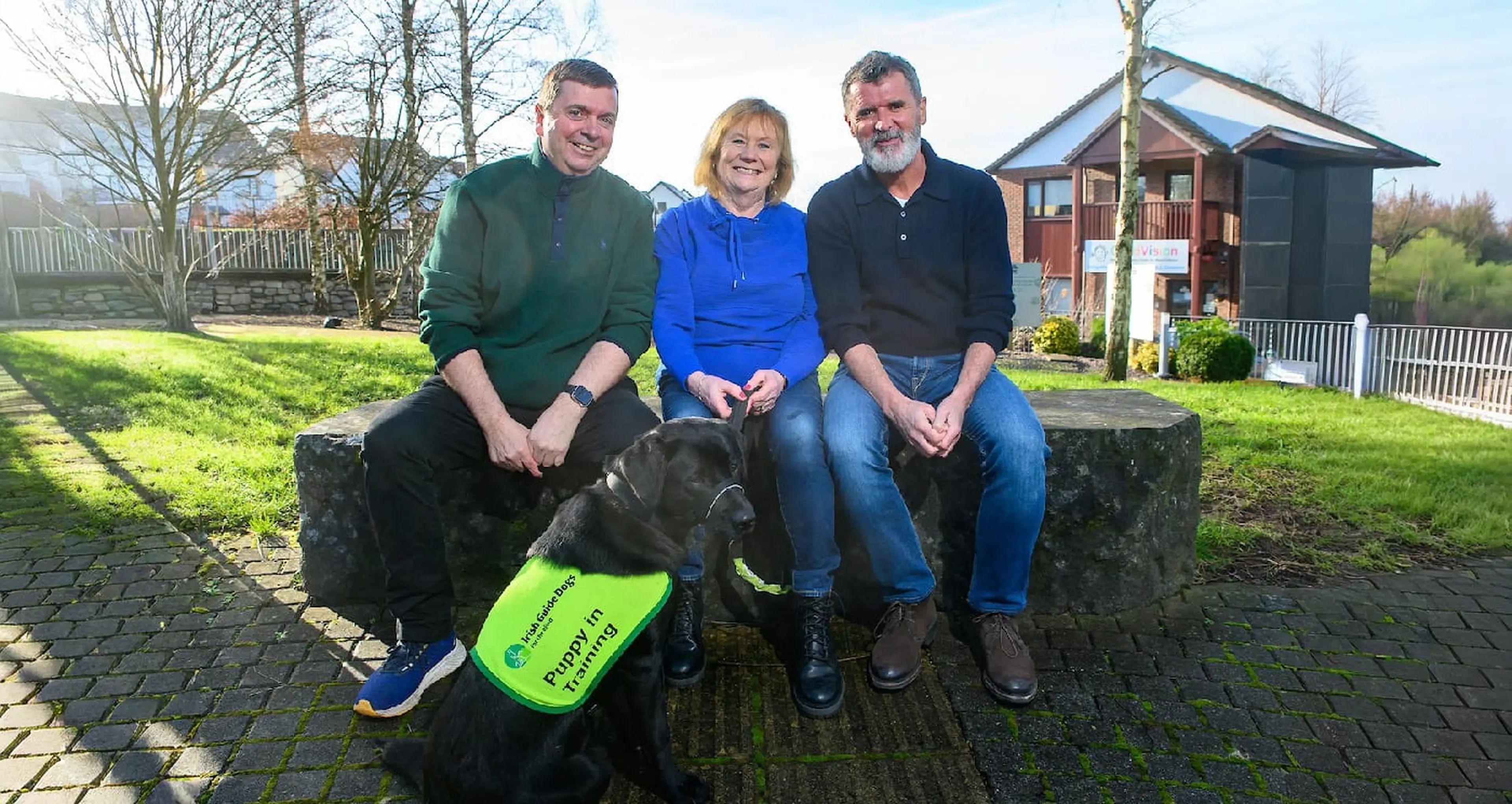 Three adults sit on a stone bench with a black service dog wearing a bright green vest in a residential garden setting.