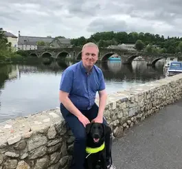 A man in a blue shirt sits on a stone wall beside a calm river with boats and buildings visible in the background.