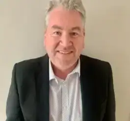 A middle-aged man with gray hair smiling at the camera while wearing a dark blazer over a light blue button-up shirt.