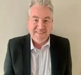 A middle-aged man with gray hair smiling at the camera while wearing a dark blazer over a light blue button-up shirt.