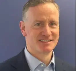 A middle-aged man with gray hair wearing a dark blazer and light blue shirt smiles at the camera against a blue background.