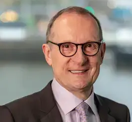 A professional headshot of a smiling middle-aged man wearing glasses, a dark blazer, and a lavender dress shirt against a blurred urban background.
