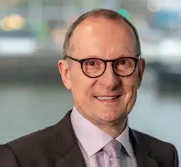 A professional headshot of a smiling middle-aged man wearing glasses, a dark blazer, and a lavender dress shirt against a blurred urban background.