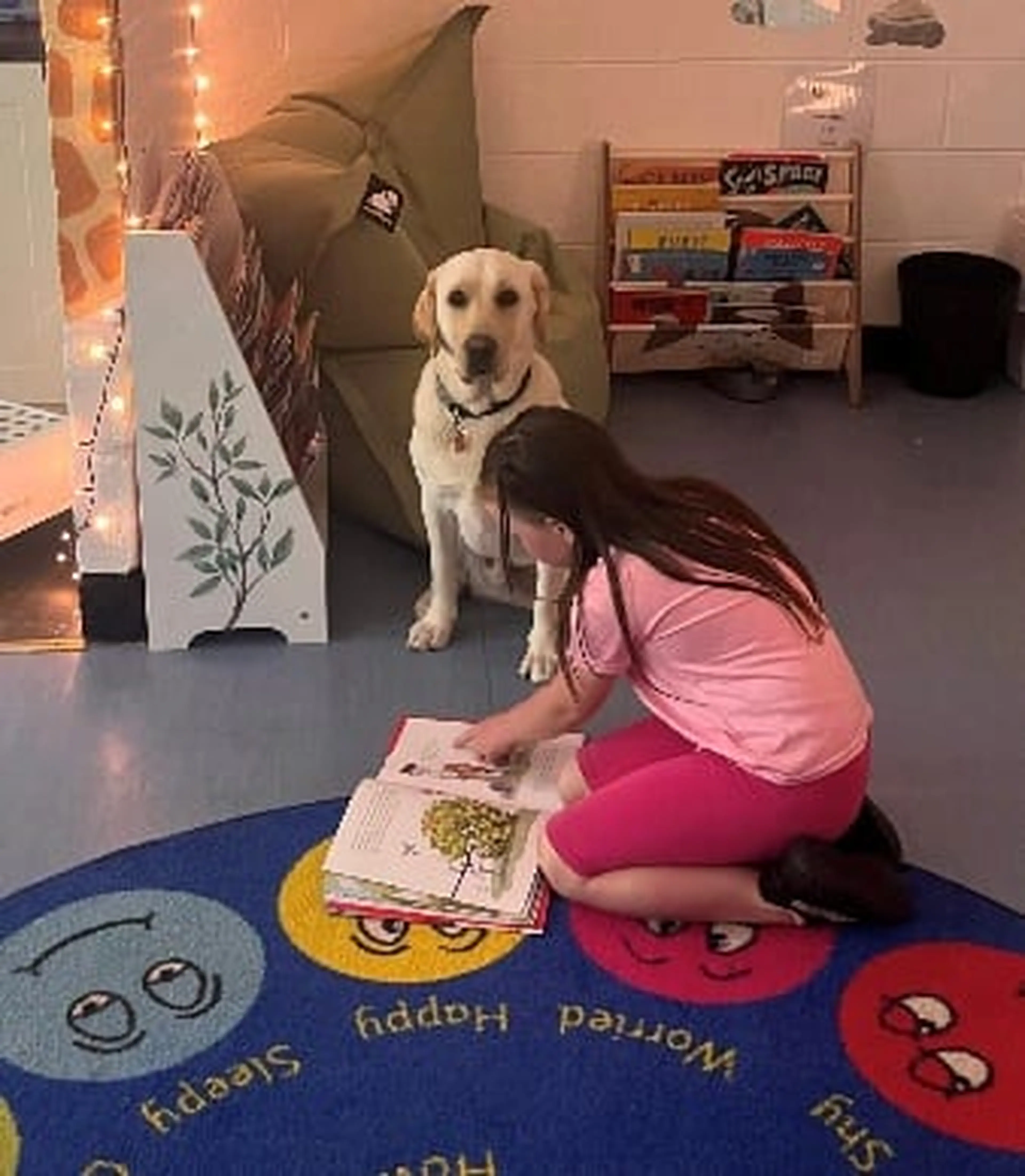 A young child in a pink shirt sits on a colorful classroom rug reading a book while a yellow Labrador retriever stands nearby watching attentively.