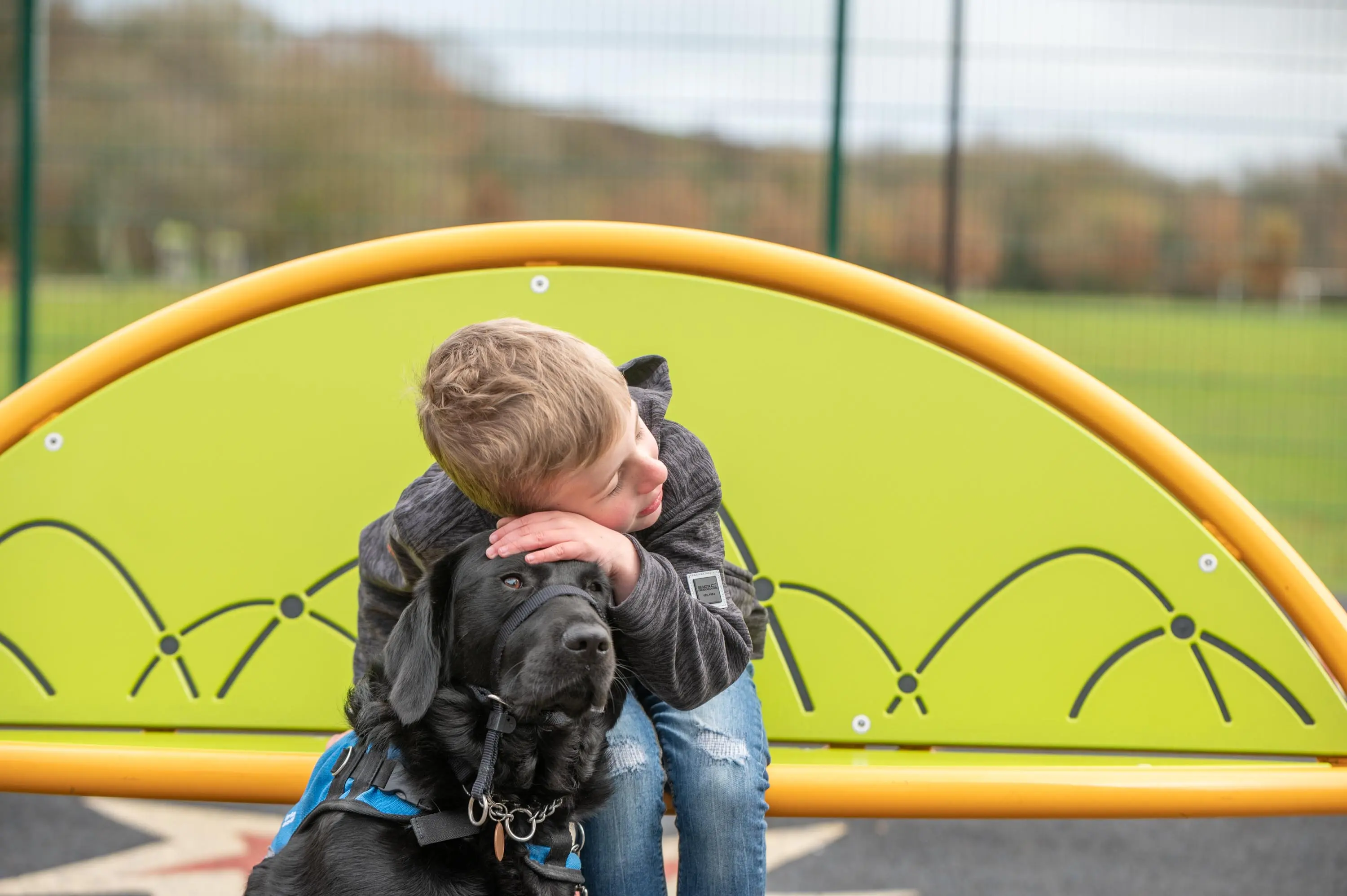 A young child sits on a black service dog in front of a bright yellow playground structure with arched design in a park setting.