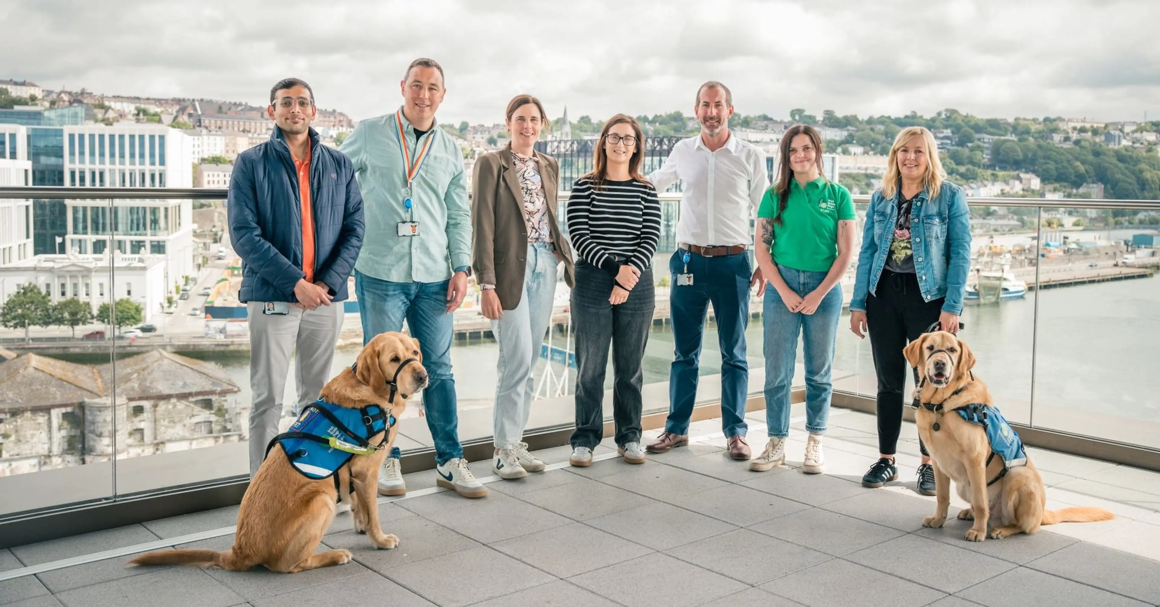 A team of seven people and two service dogs stand together on a rooftop terrace overlooking a riverside city with buildings and green spaces in the background.