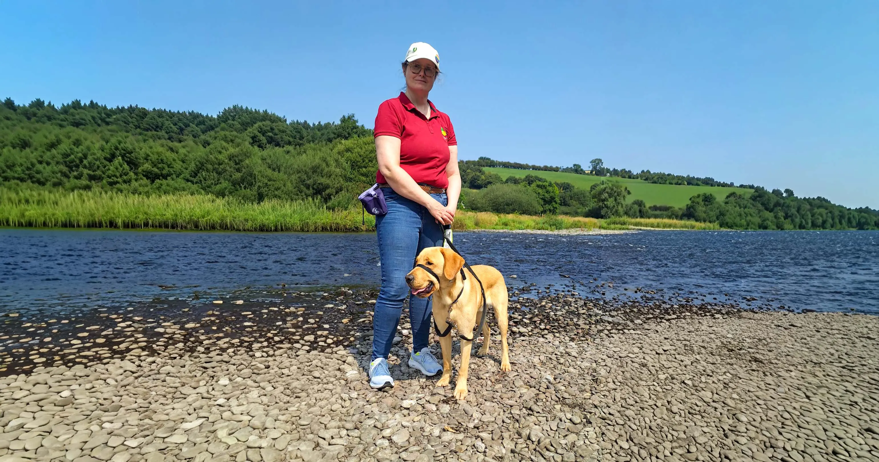 A woman in a red polo shirt and white cap stands on a pebbly beach with a yellow Labrador Retriever on a leash, with a calm lake and forested hillside in the background under clear
