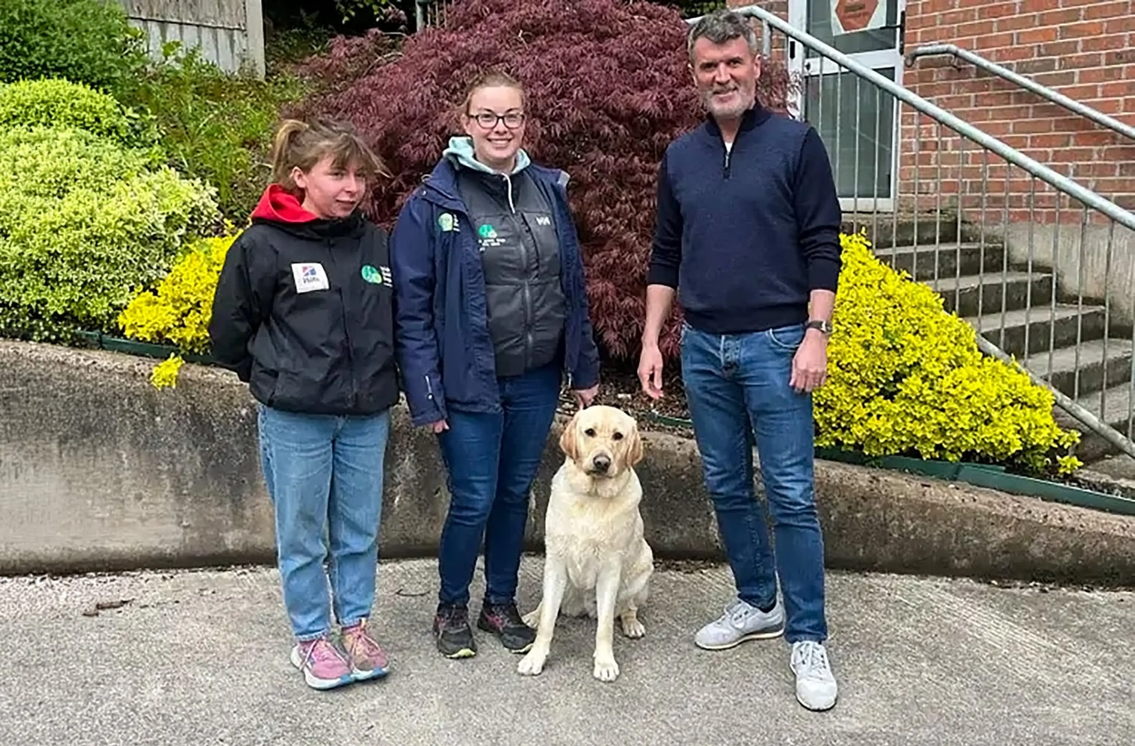 A family of three stands with their cream-colored Labrador Retriever in front of a brick building surrounded by yellow and burgundy foliage.