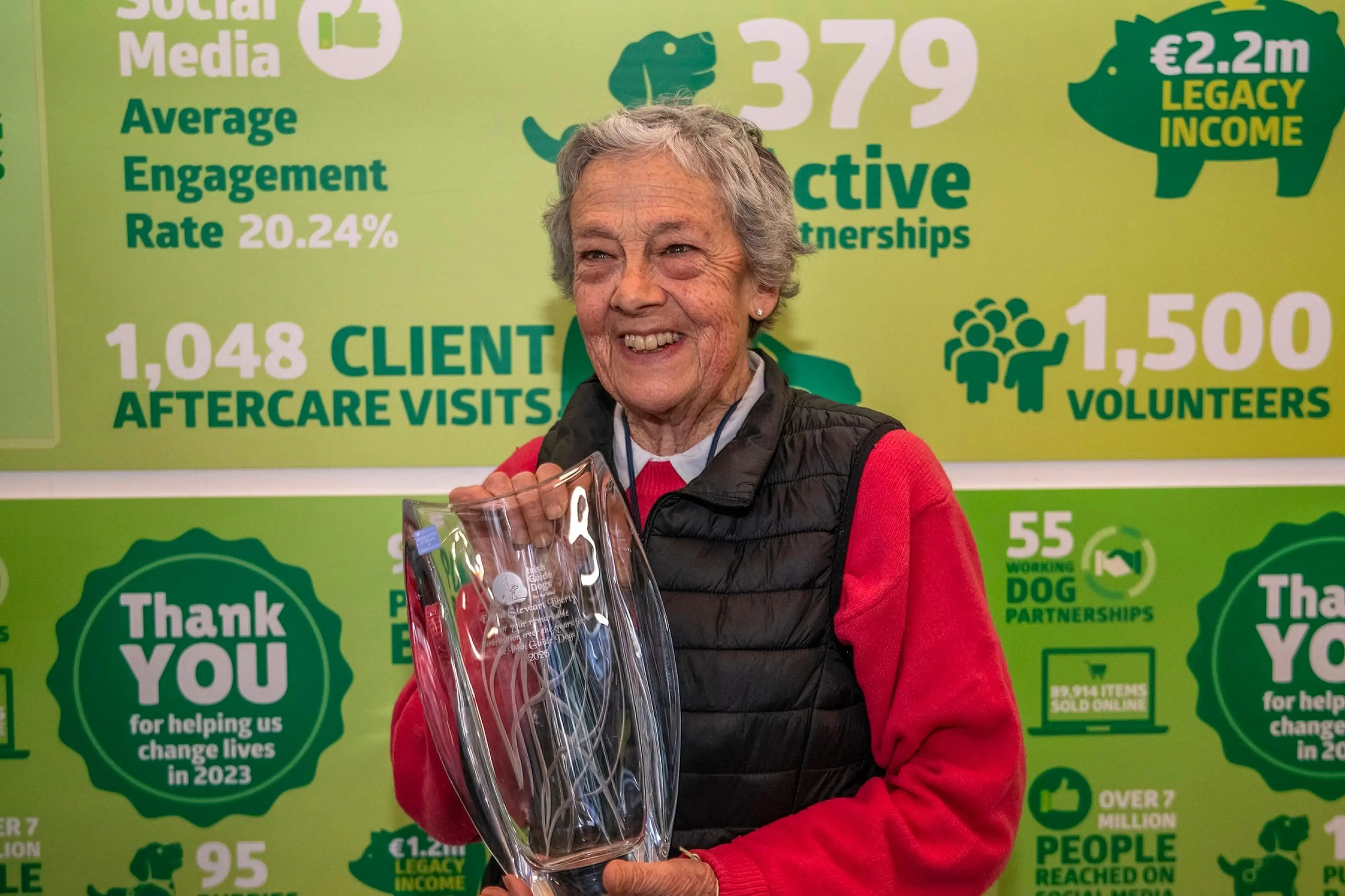 The image shows a smiling elderly woman holding a glass container, standing in front of a colorful poster displaying statistics and information about a charitable organization.