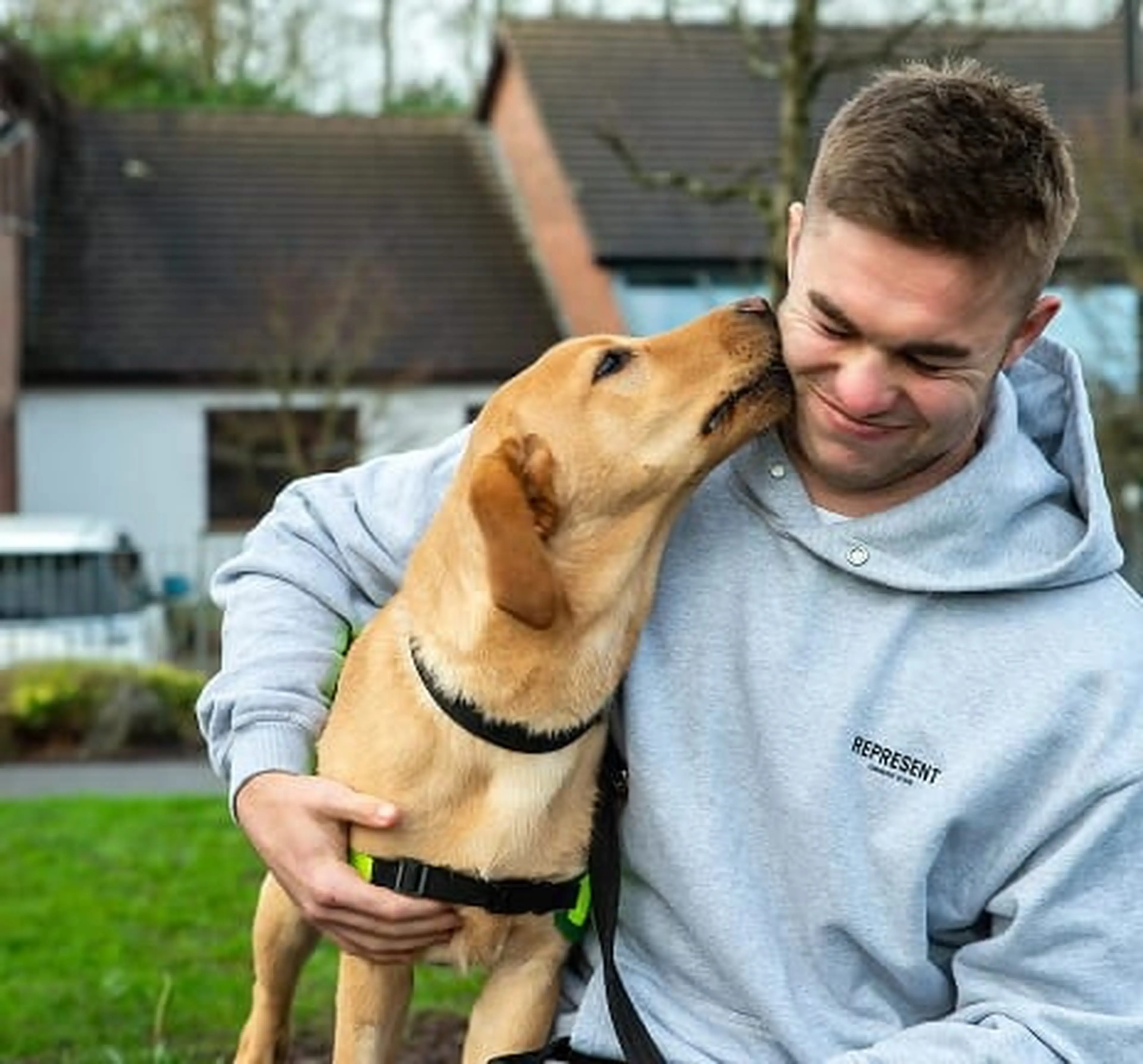 A young man in a gray hoodie smiles while holding a brown dog that is nuzzling his face in a residential backyard.