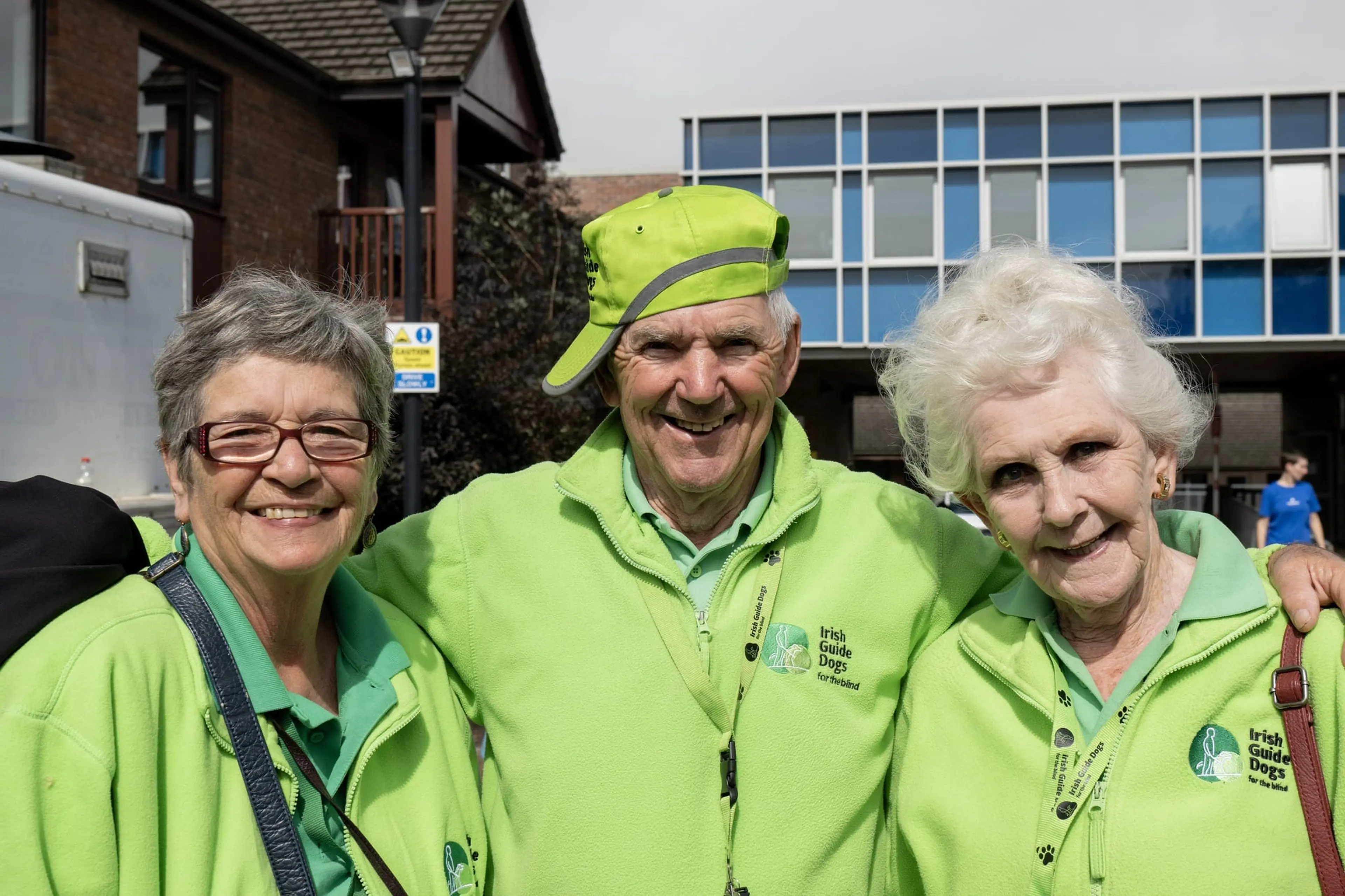 Three smiling elderly people in green jackets, one wearing a matching cap, stand together outside a building on a cloudy day.