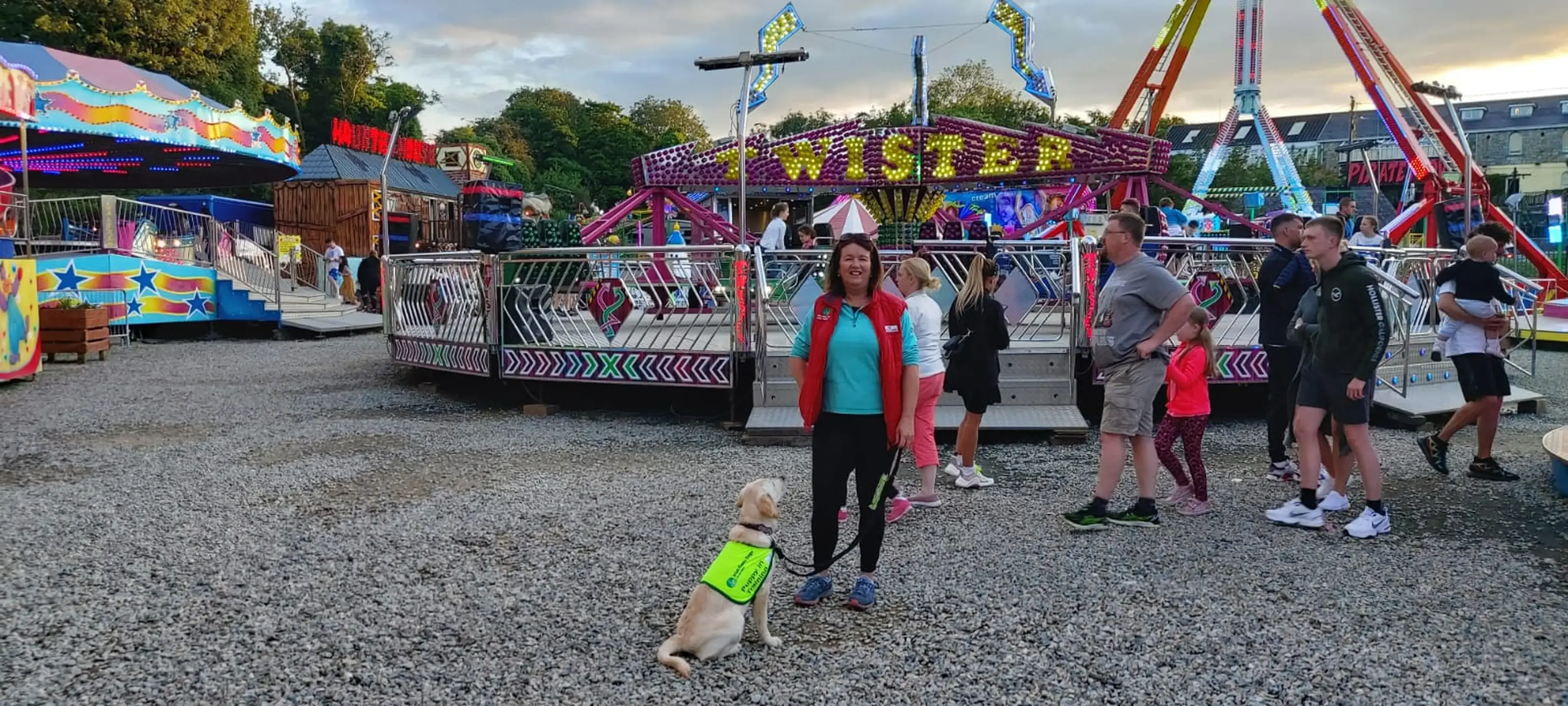 A small light-colored dog wearing a neon yellow vest sits on gravel at a colorful fair or carnival, surrounded by visitors and vibrant rides in the background.