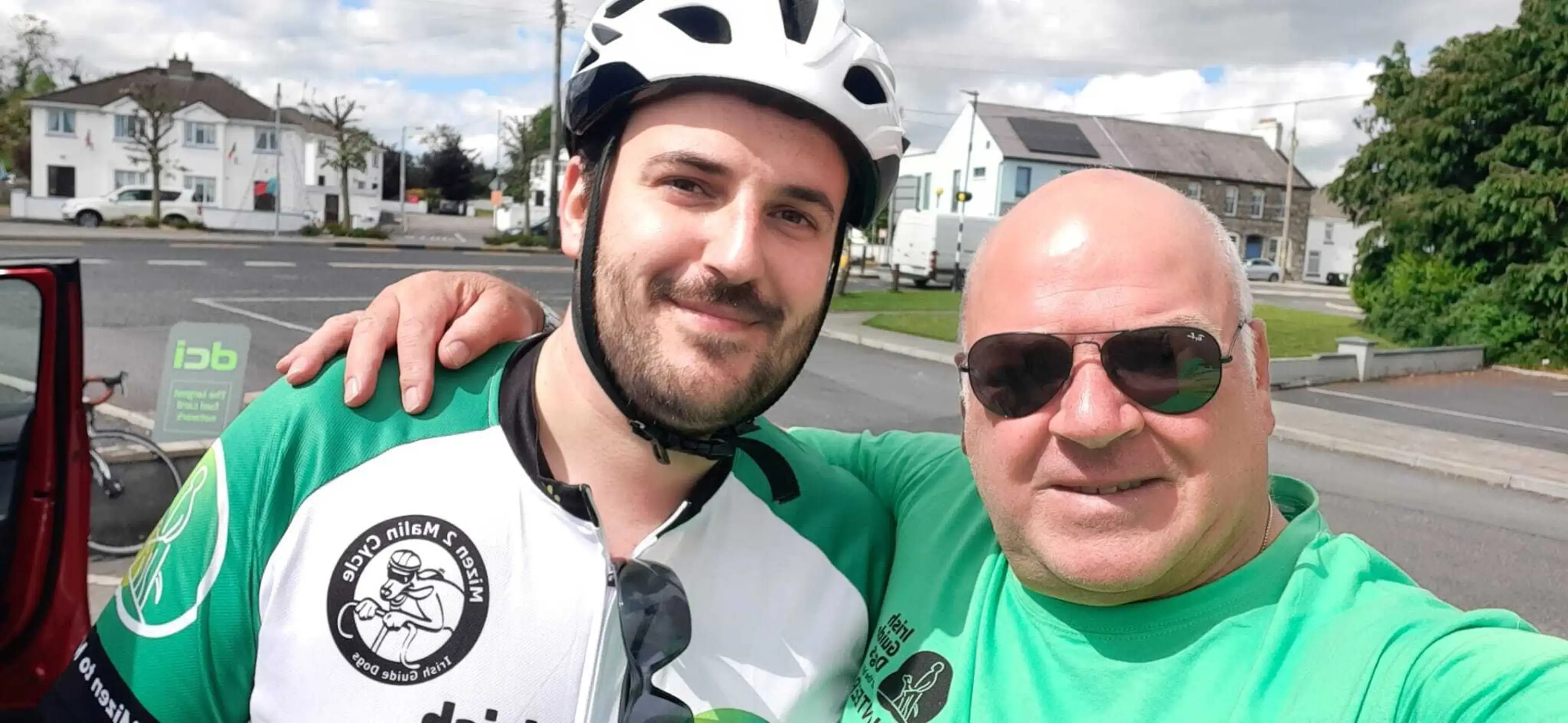 Two men in green cycling gear pose together on a residential street, with one wearing a white helmet and the other wearing sunglasses.