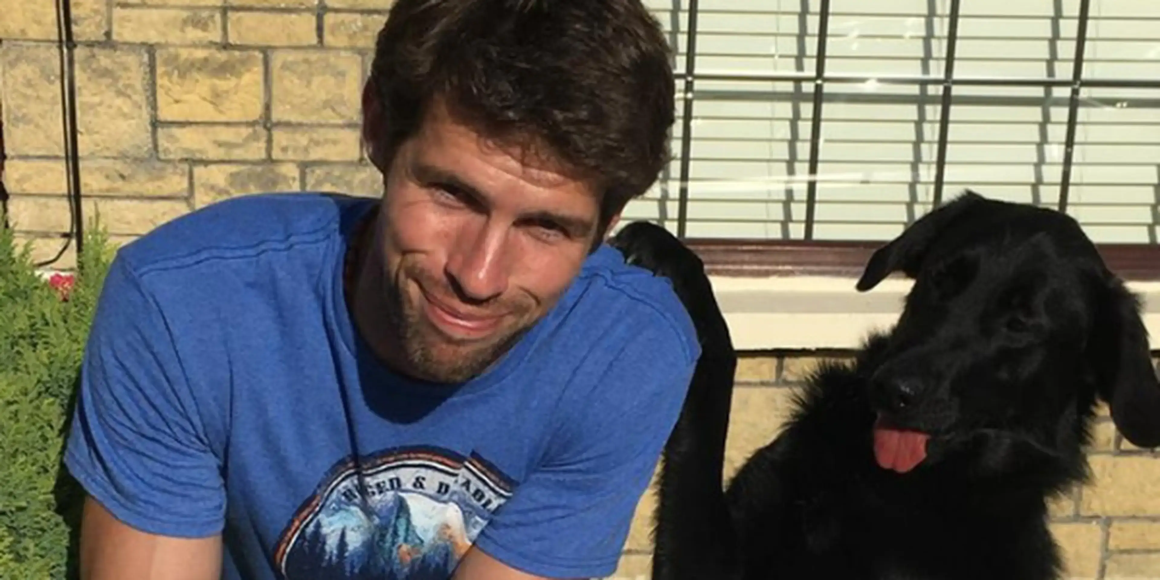 A man in a blue graphic t-shirt smiles while interacting with a black dog wearing a red collar in a brick-walled outdoor patio setting.