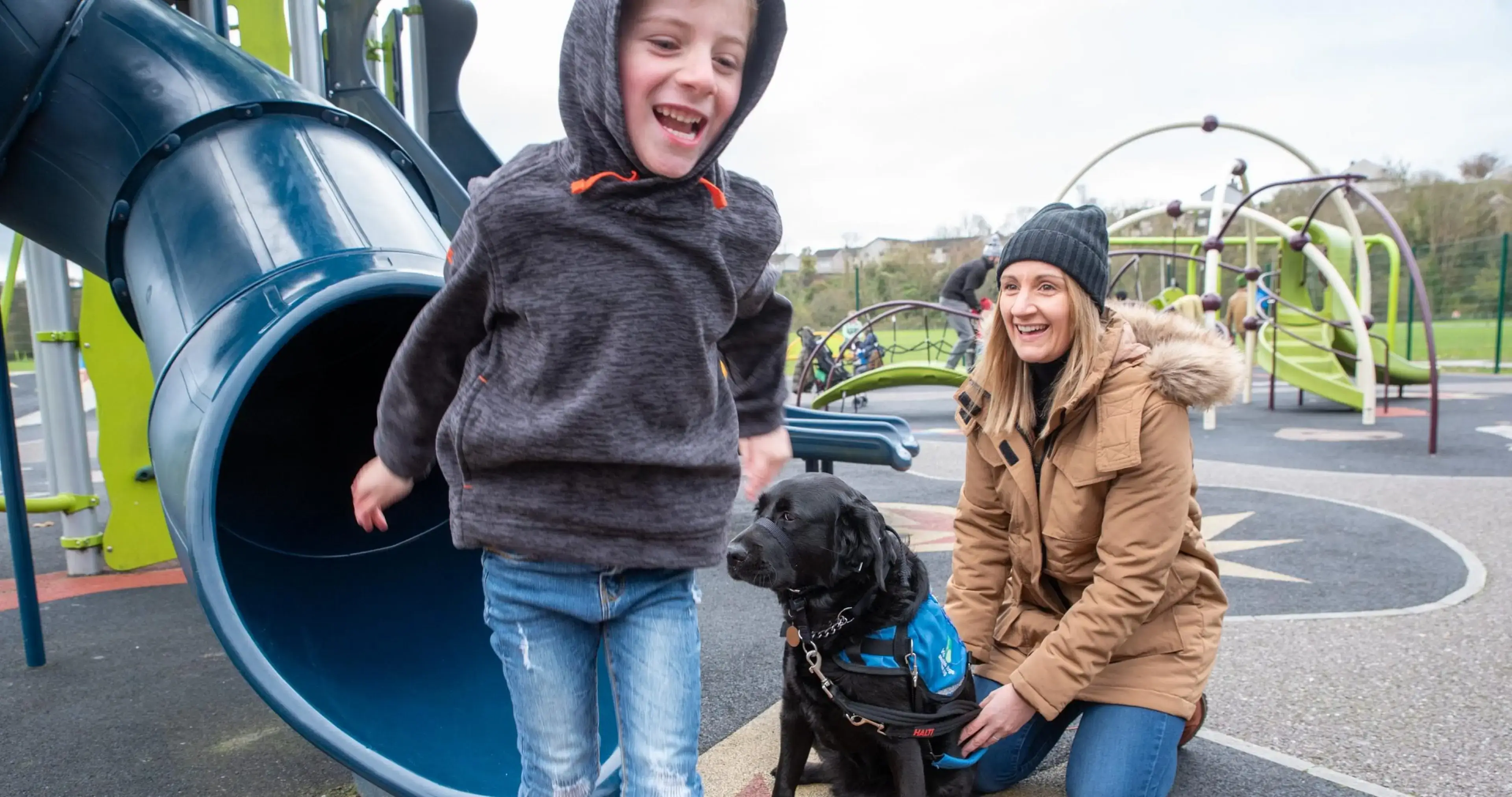 A woman and child laugh together at a playground with a black service dog wearing a blue vest beside them, with a blue tunnel slide and climbing structures visible in the backgroun