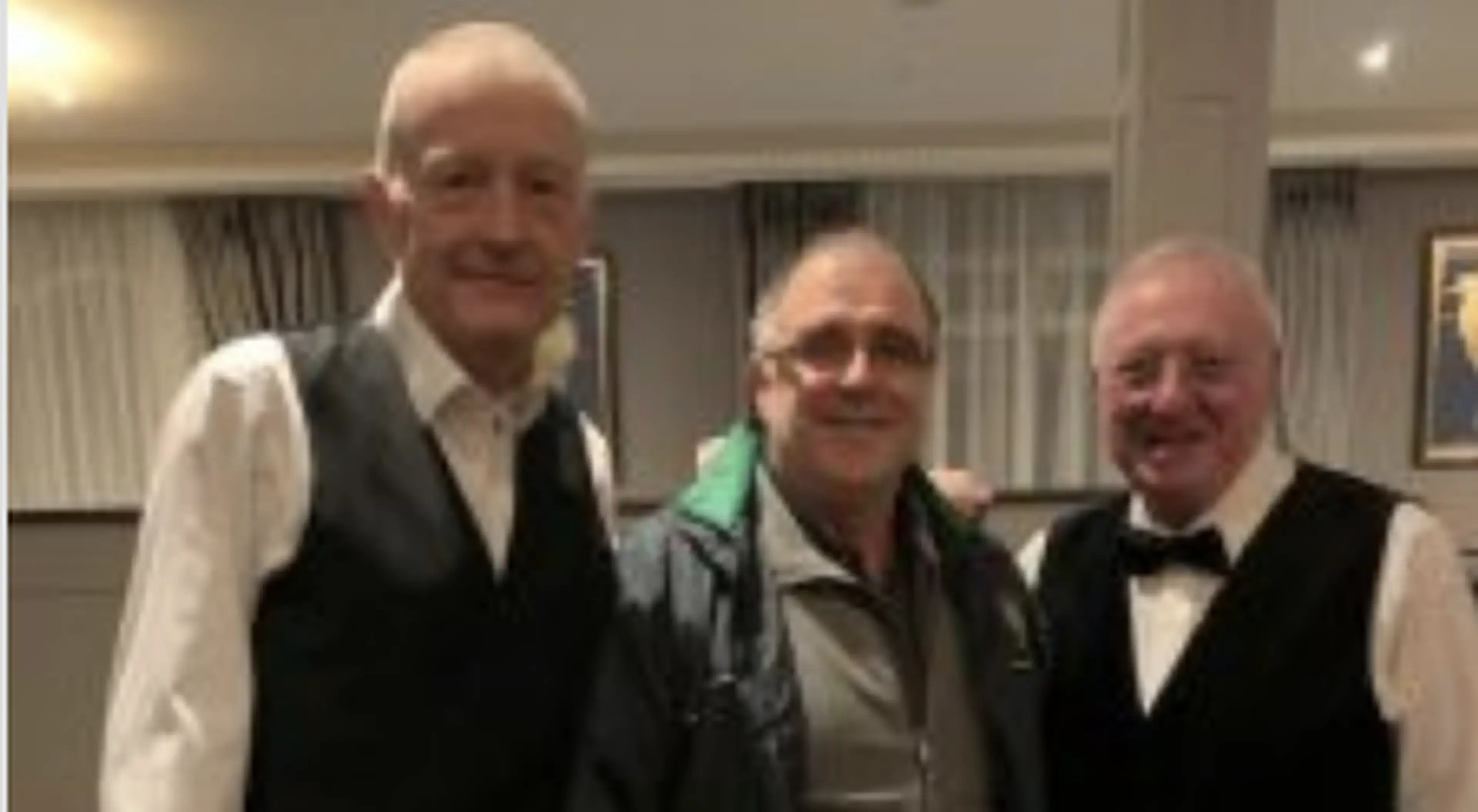 Three older men pose together for a photo at what appears to be a formal indoor event, with two wearing vests and one in a green shirt.
