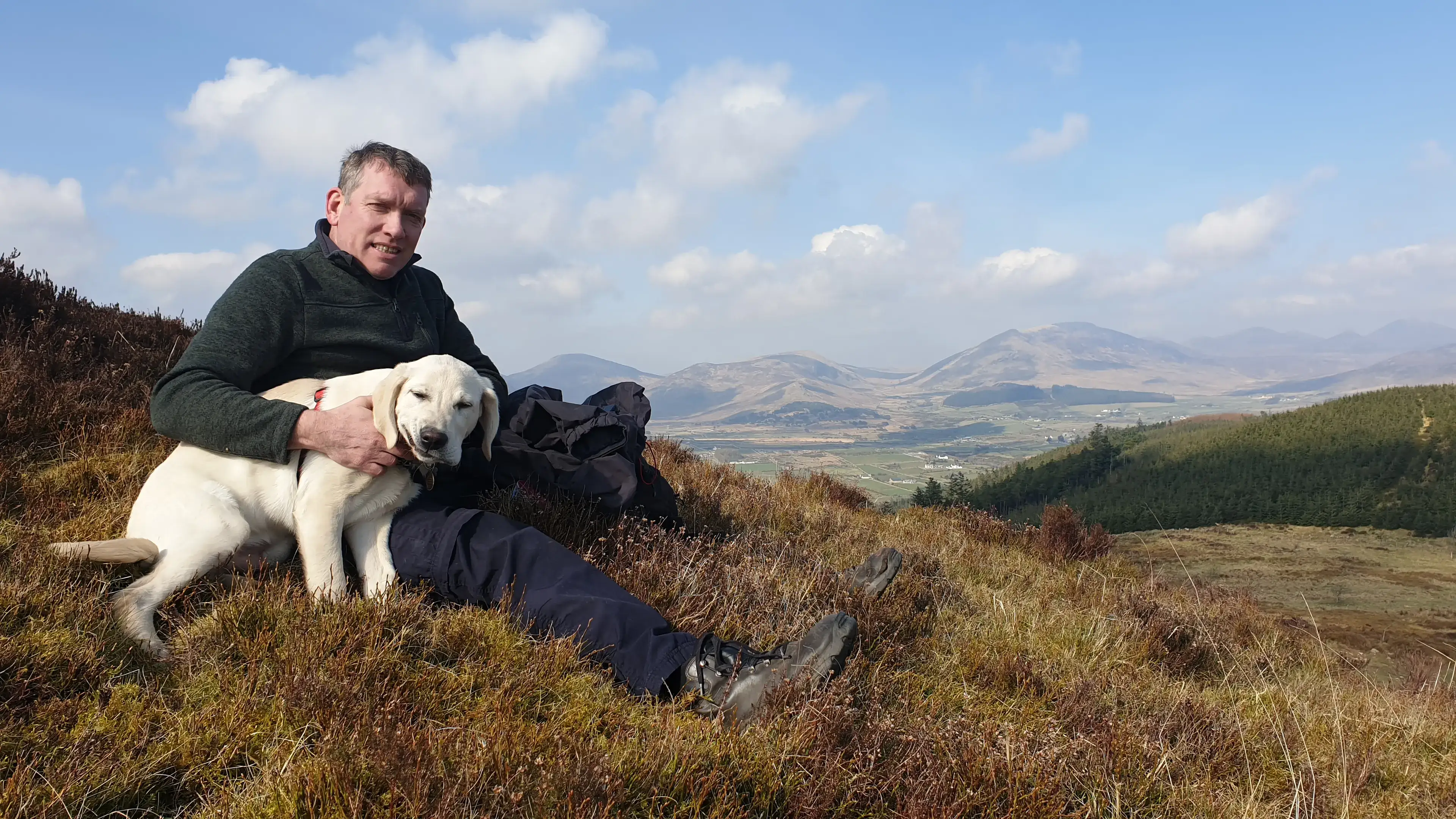 A man in a dark jacket sits on a hillside with a white Labrador dog, overlooking a valley with mountains in the distance under a blue sky with white clouds.
