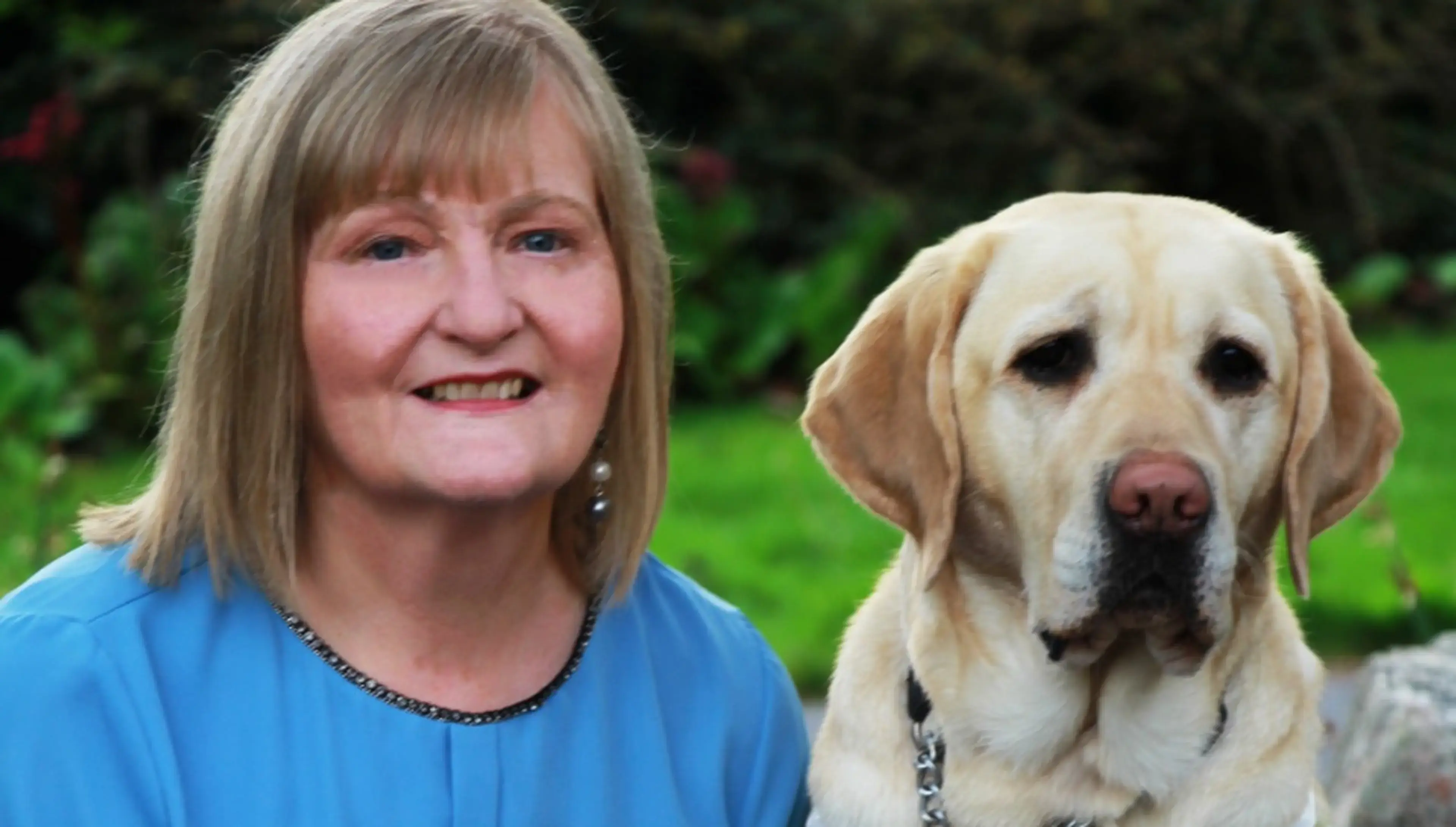 A smiling woman in a blue shirt poses with a yellow Labrador Retriever in a garden setting.