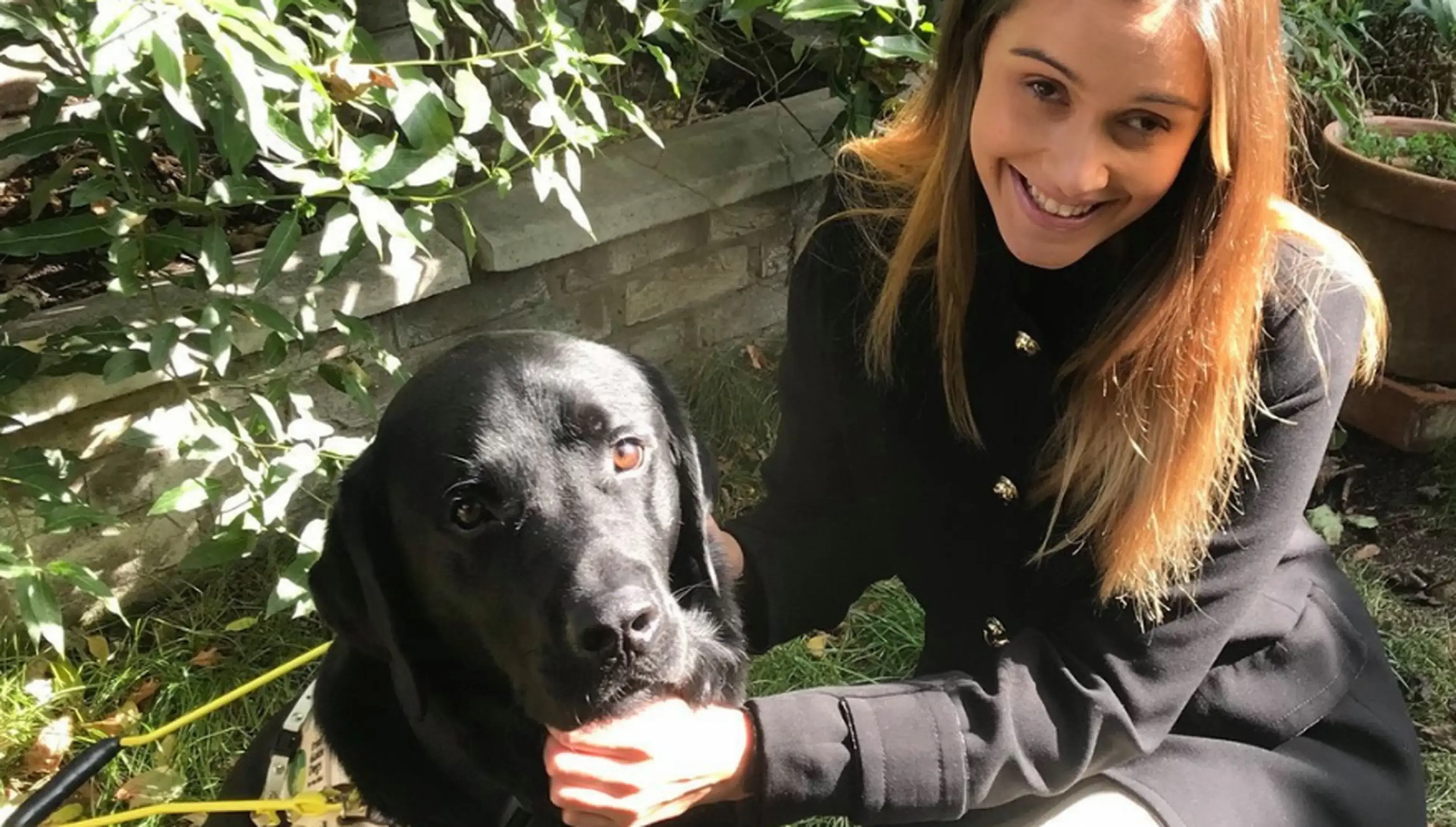 A smiling young woman with reddish-brown hair sits outdoors next to a black dog wearing a reflective collar, with green foliage visible in the background.