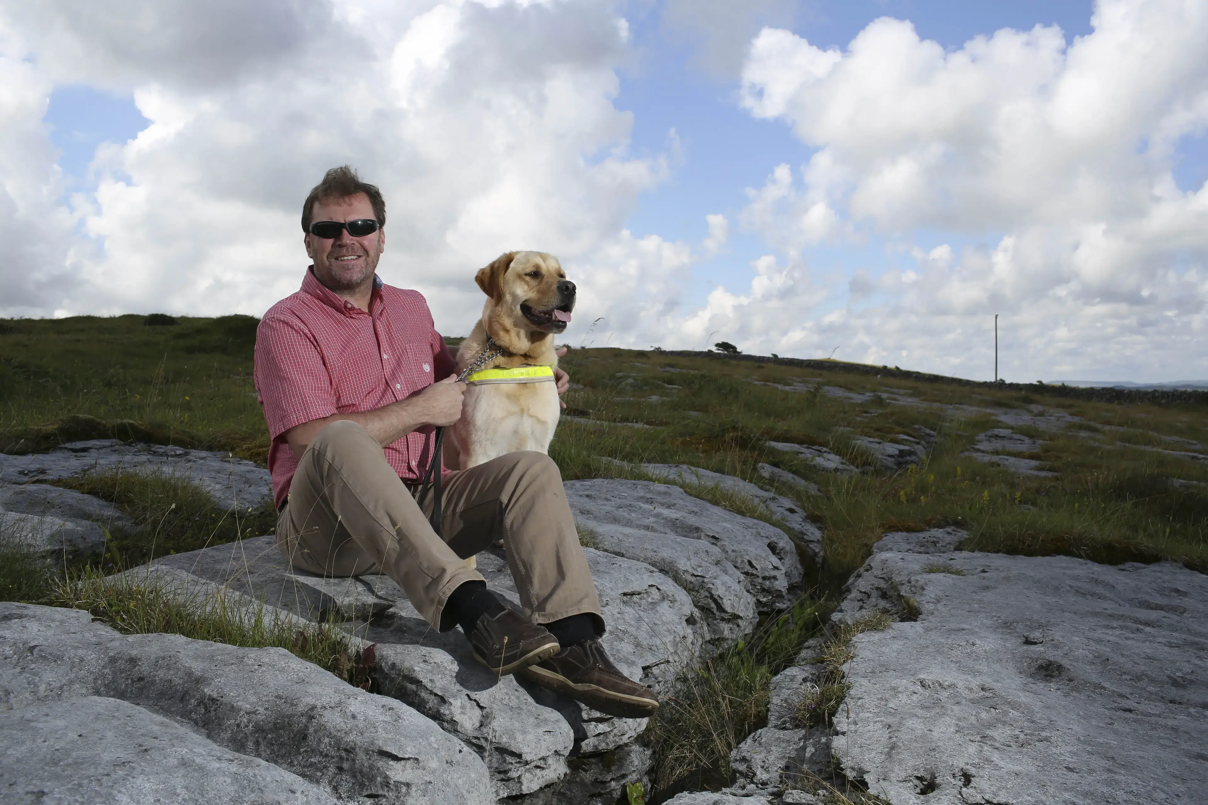 A man wearing sunglasses and a pink shirt crouches beside a yellow Labrador Retriever on rocky terrain with rolling hills and cloudy sky in the background.