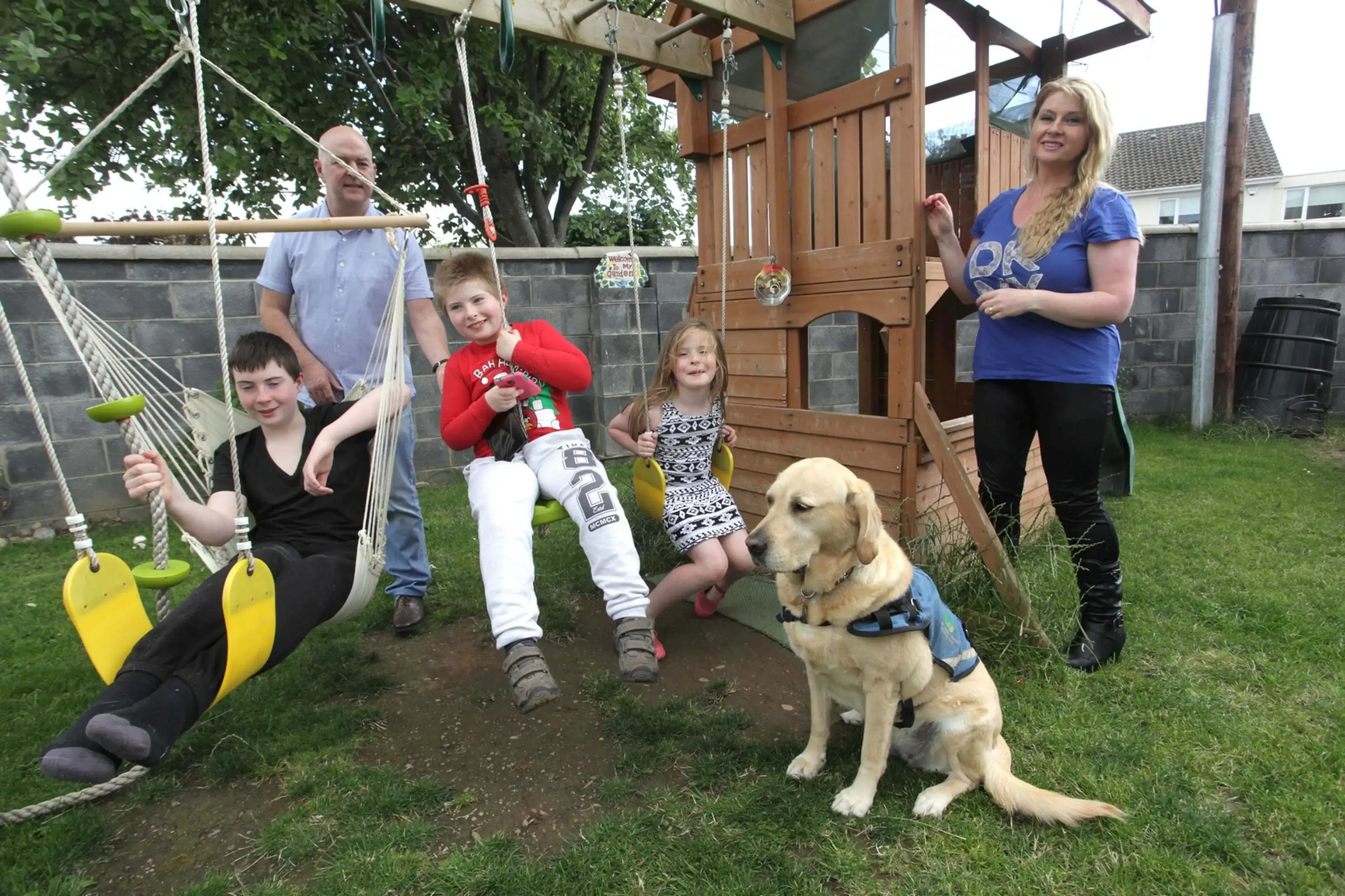 A family of five and a yellow Labrador dog pose together in a backyard with a wooden playset and swings.