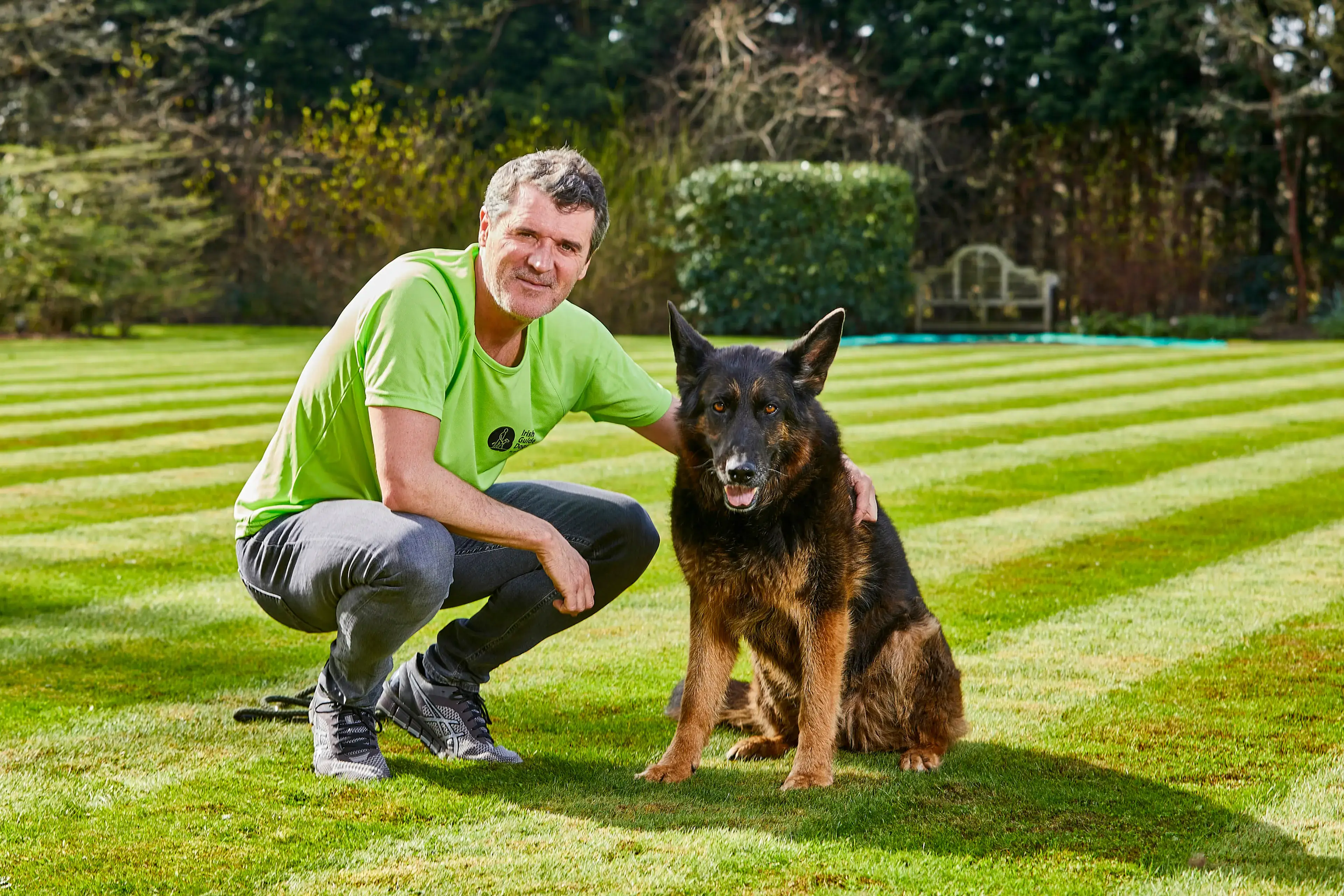 A man in a lime green t-shirt crouches beside a black and tan German Shepherd on a striped lawn, with trees and a garden bench visible in the background.