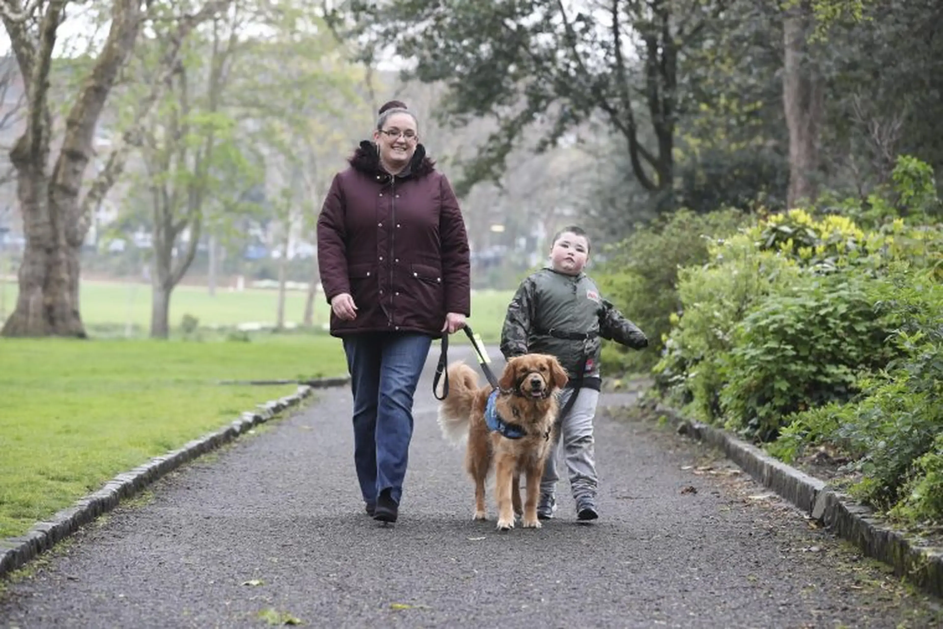 A woman and child walk a brown dog on a leash along a tree-lined park path on an overcast day.