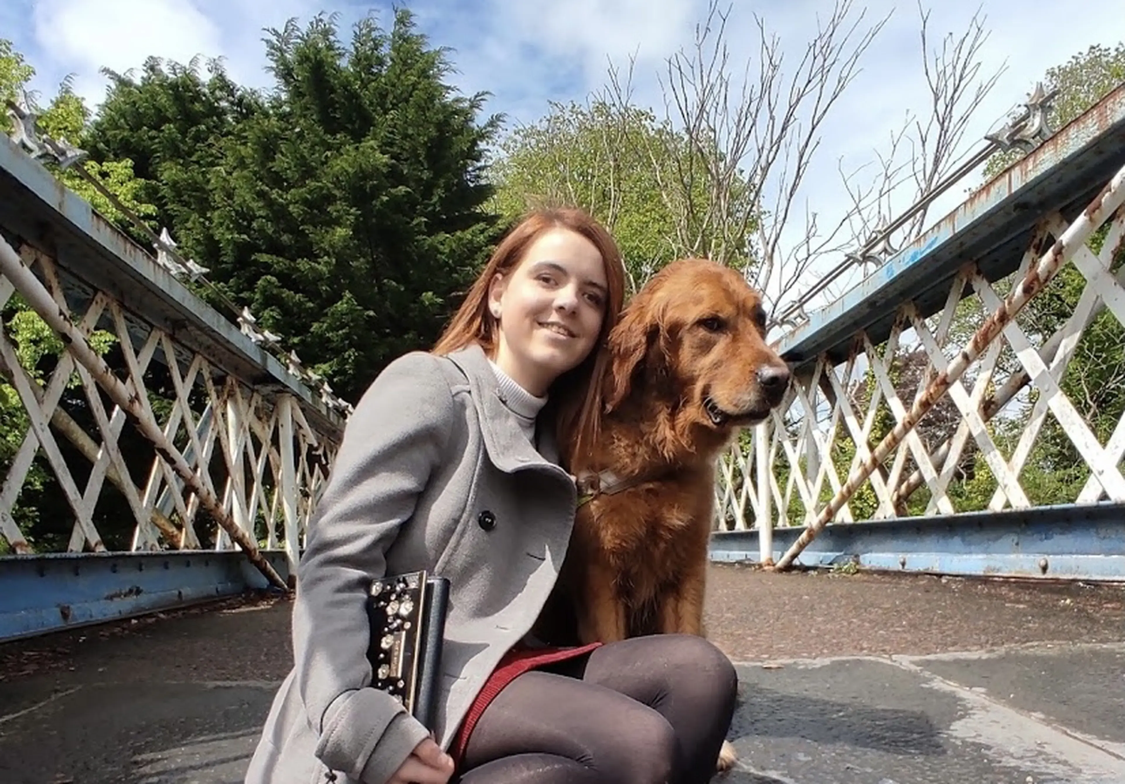 A smiling young woman with reddish-brown hair crouches beside a golden-brown dog on a bridge, holding a guitar while posing for the camera.