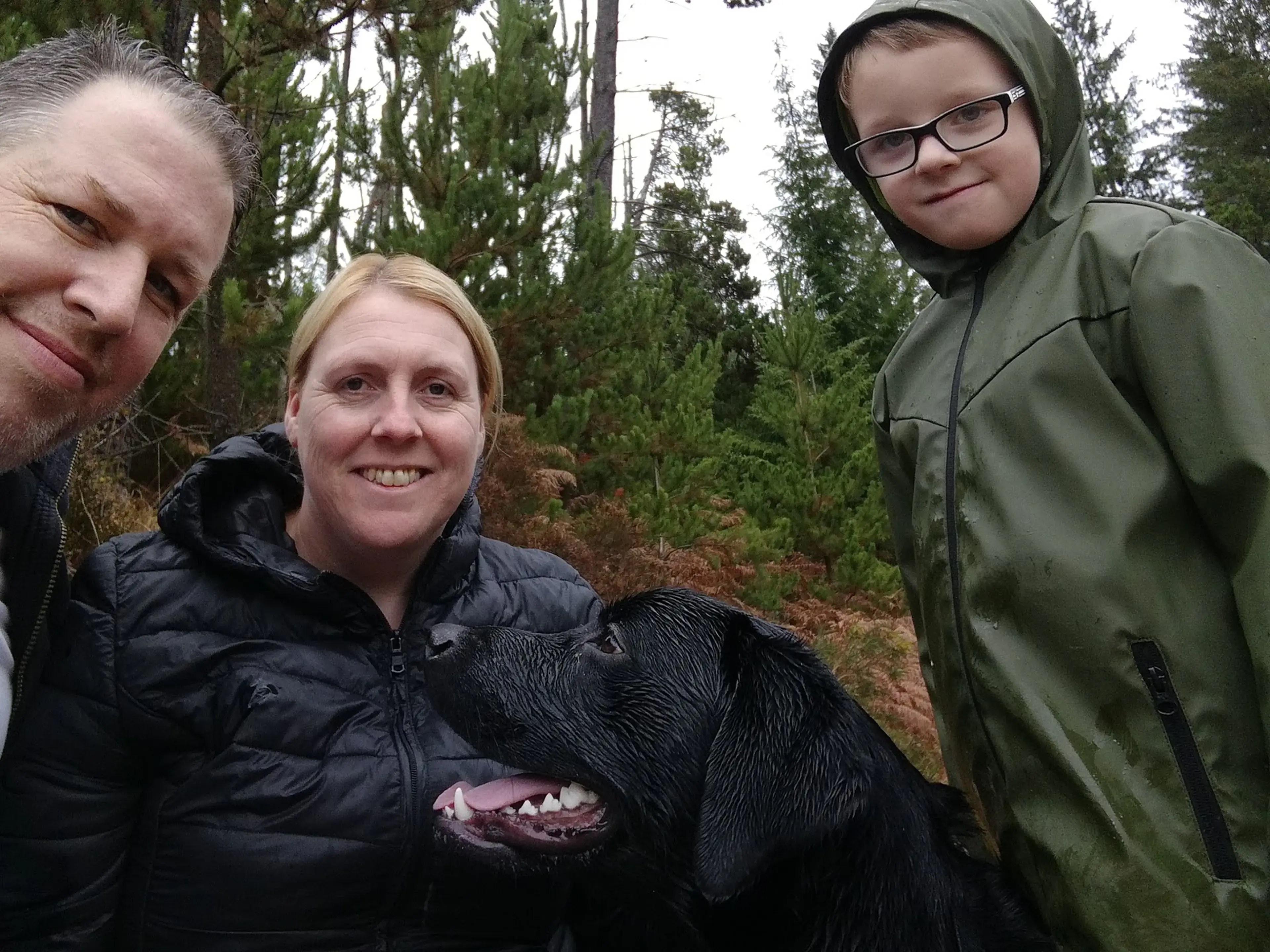Three people pose with a black dog in a forested area with evergreen trees in the background.