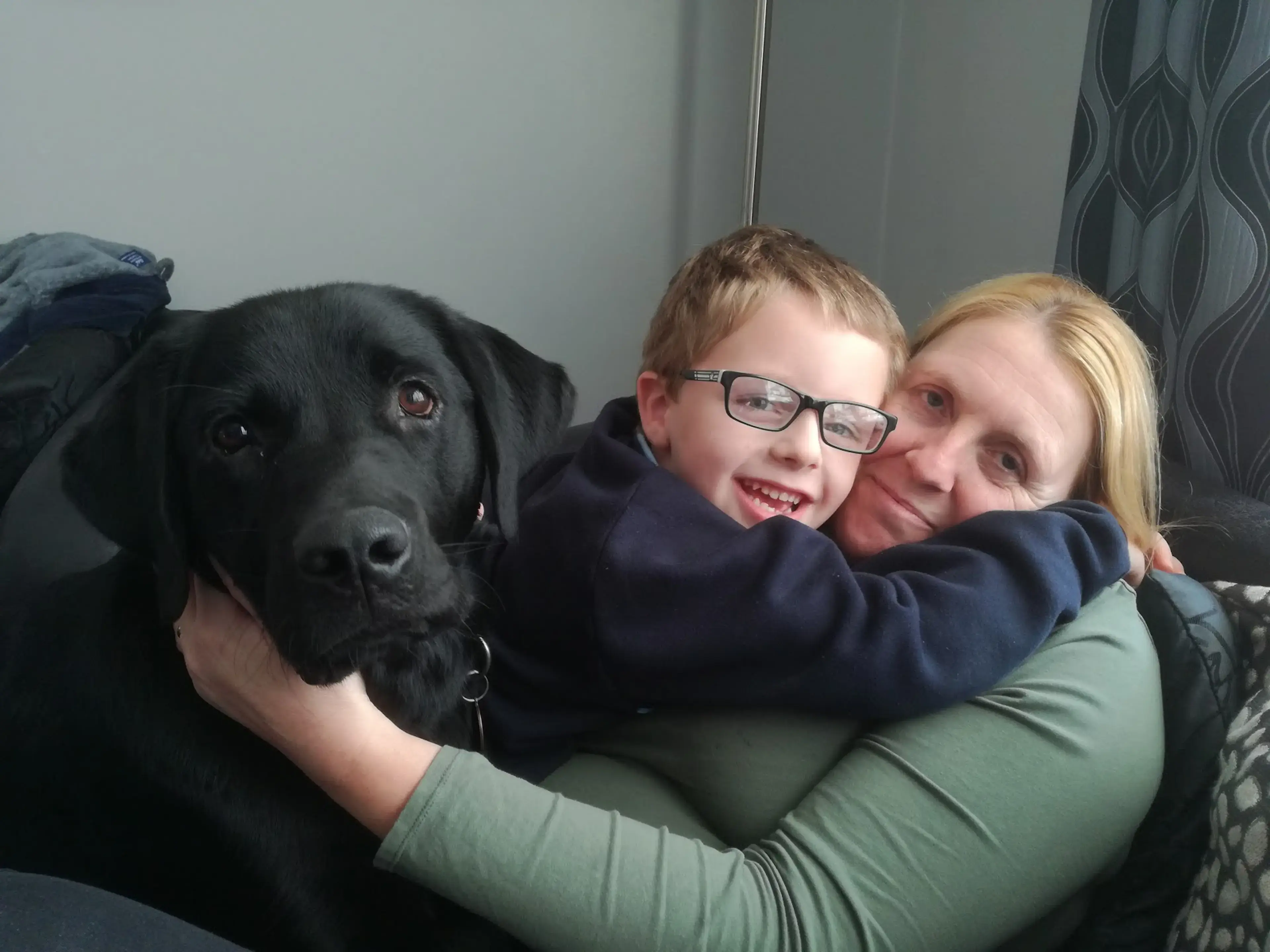A woman and young boy wearing glasses smile while posing with a black Labrador dog on a couch indoors.