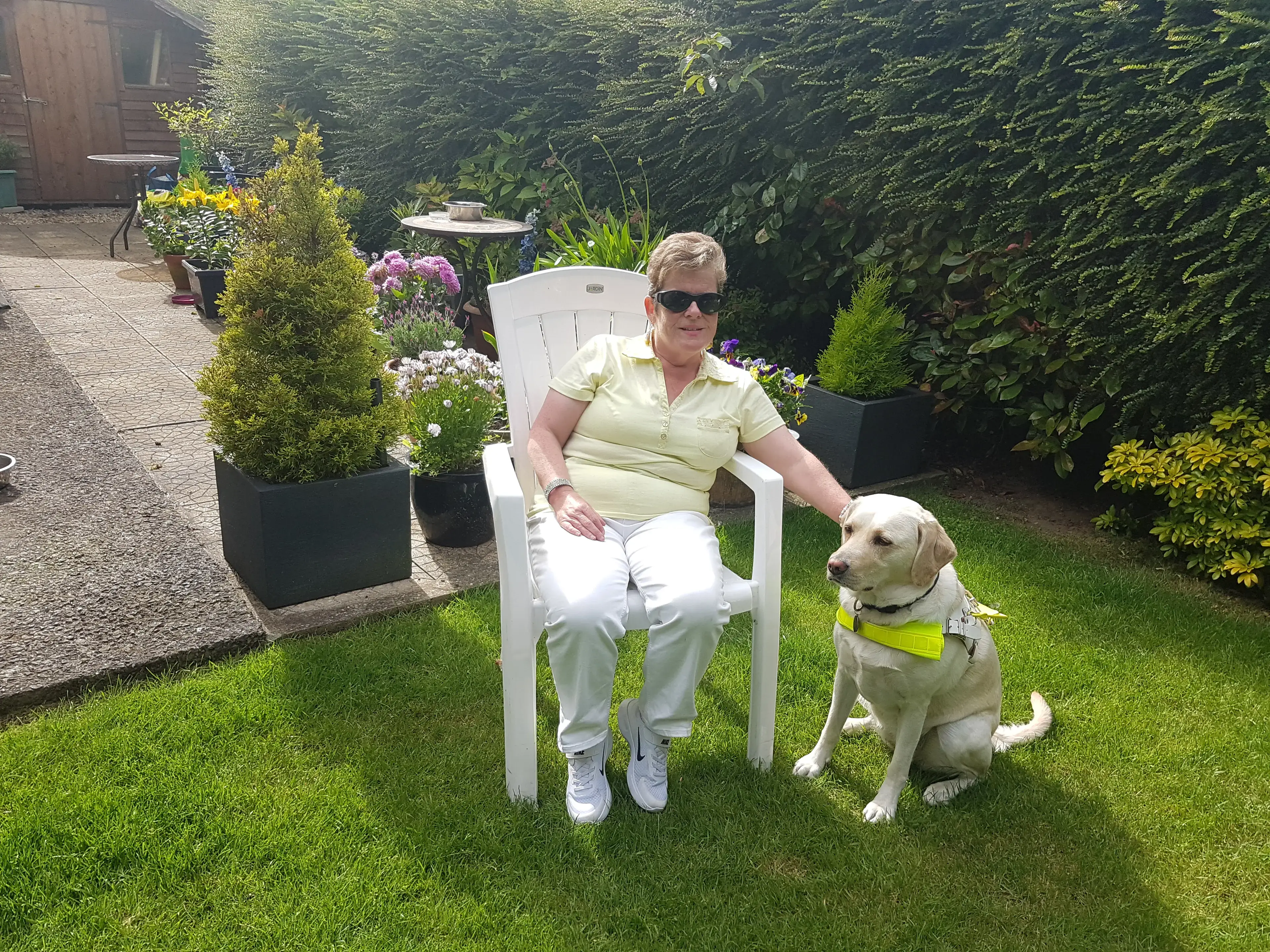 A woman in a yellow shirt and sunglasses sits in a white garden chair next to her service dog, a yellow Labrador wearing a yellow vest, on a sunny lawn surrounded by flowering plan