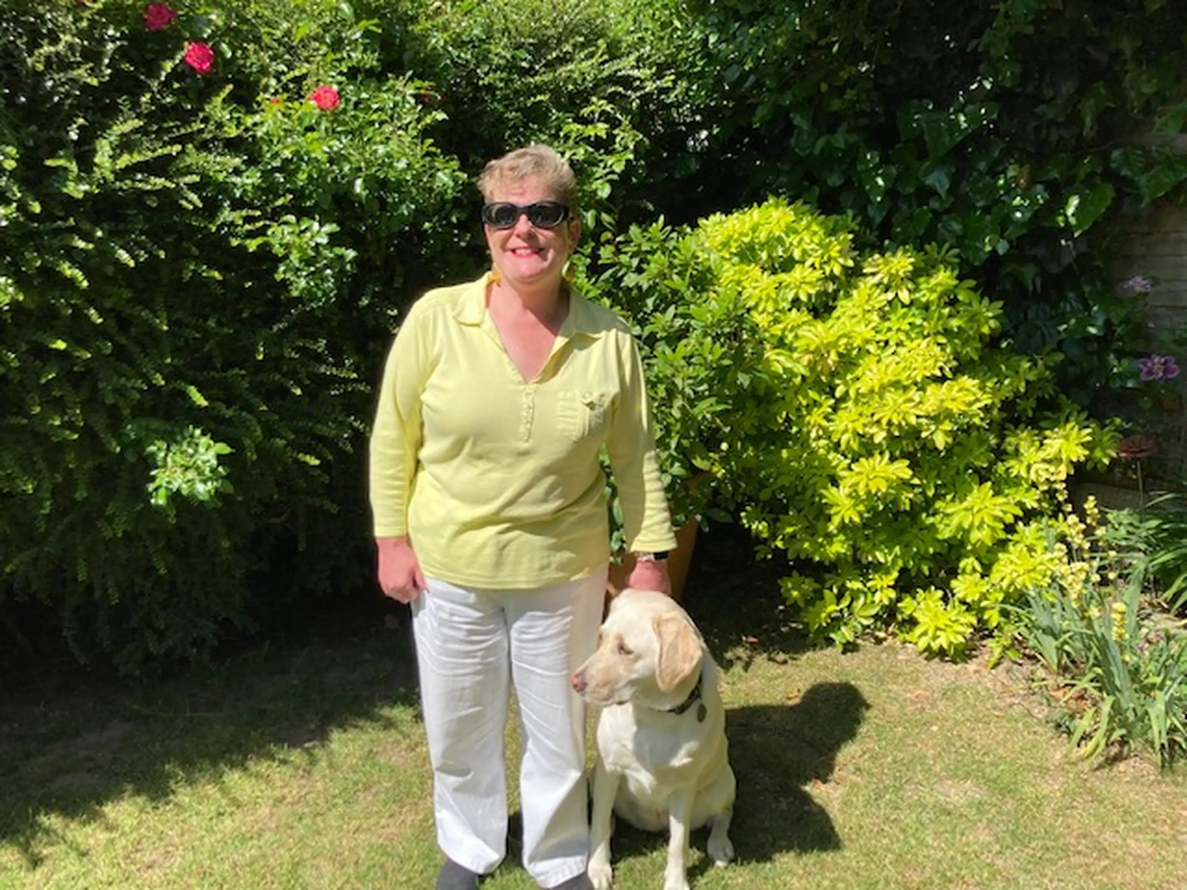 A woman wearing a yellow shirt and sunglasses stands in a sunny garden with a light-colored dog at her side, surrounded by green foliage and flowering plants.