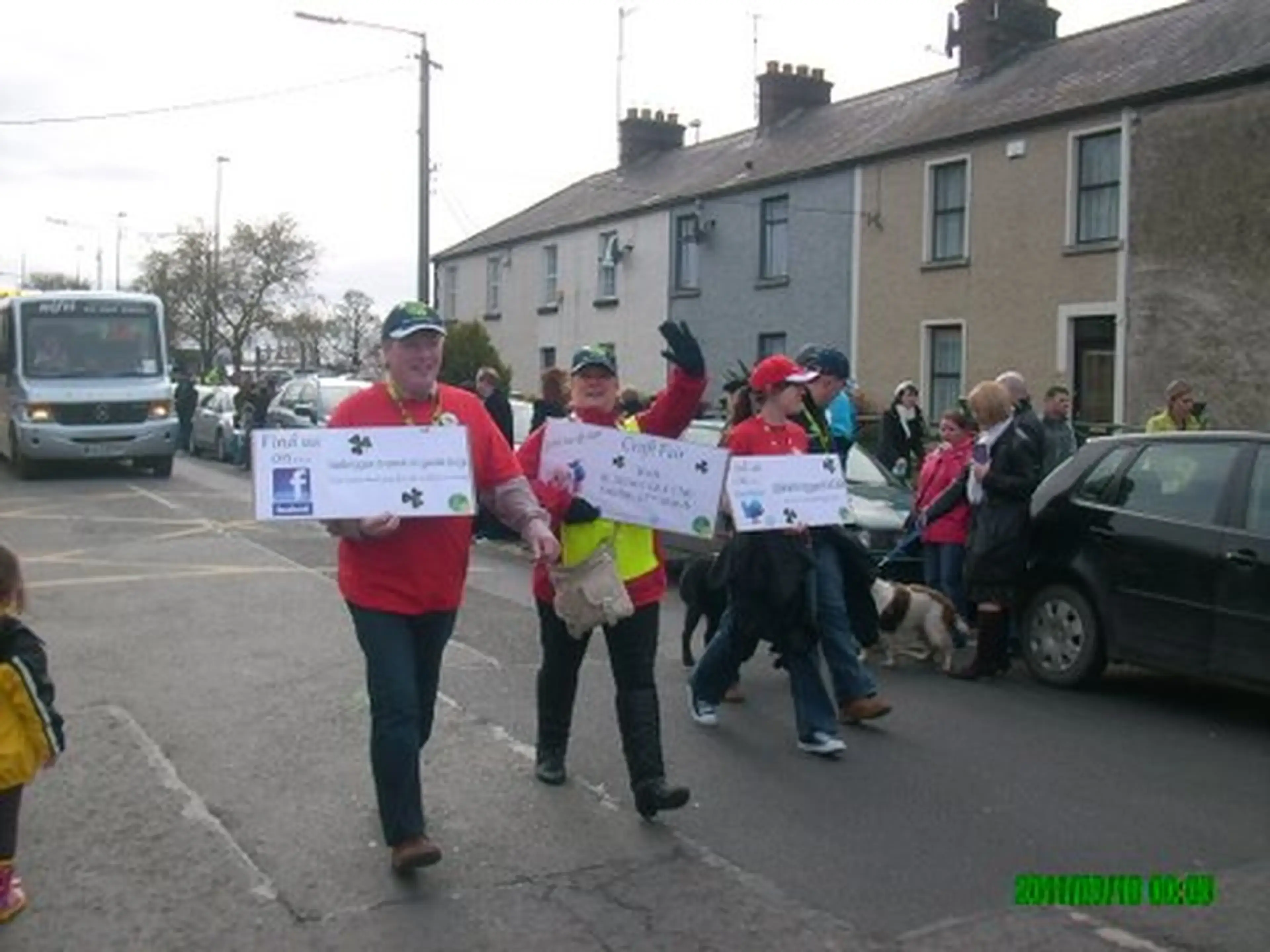 A group of people wearing red shirts and caps participate in a street parade or charity walk, holding large commemorative checks, with row houses and parked cars visible in the bac
