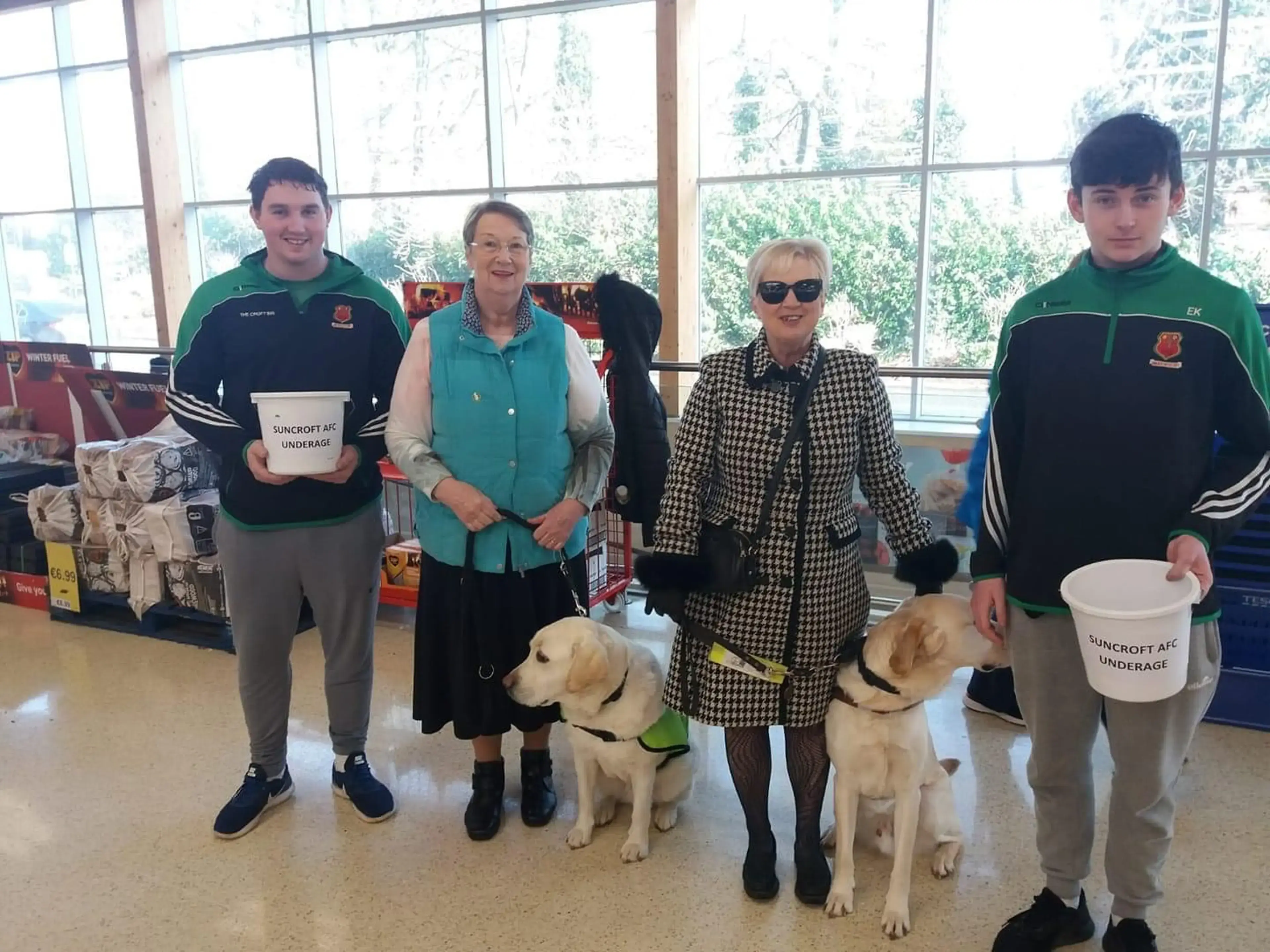 Five people, including two young men in green and black sports jackets and two women, stand indoors holding collection buckets for Suncroft AFC Underage while posing with two yello