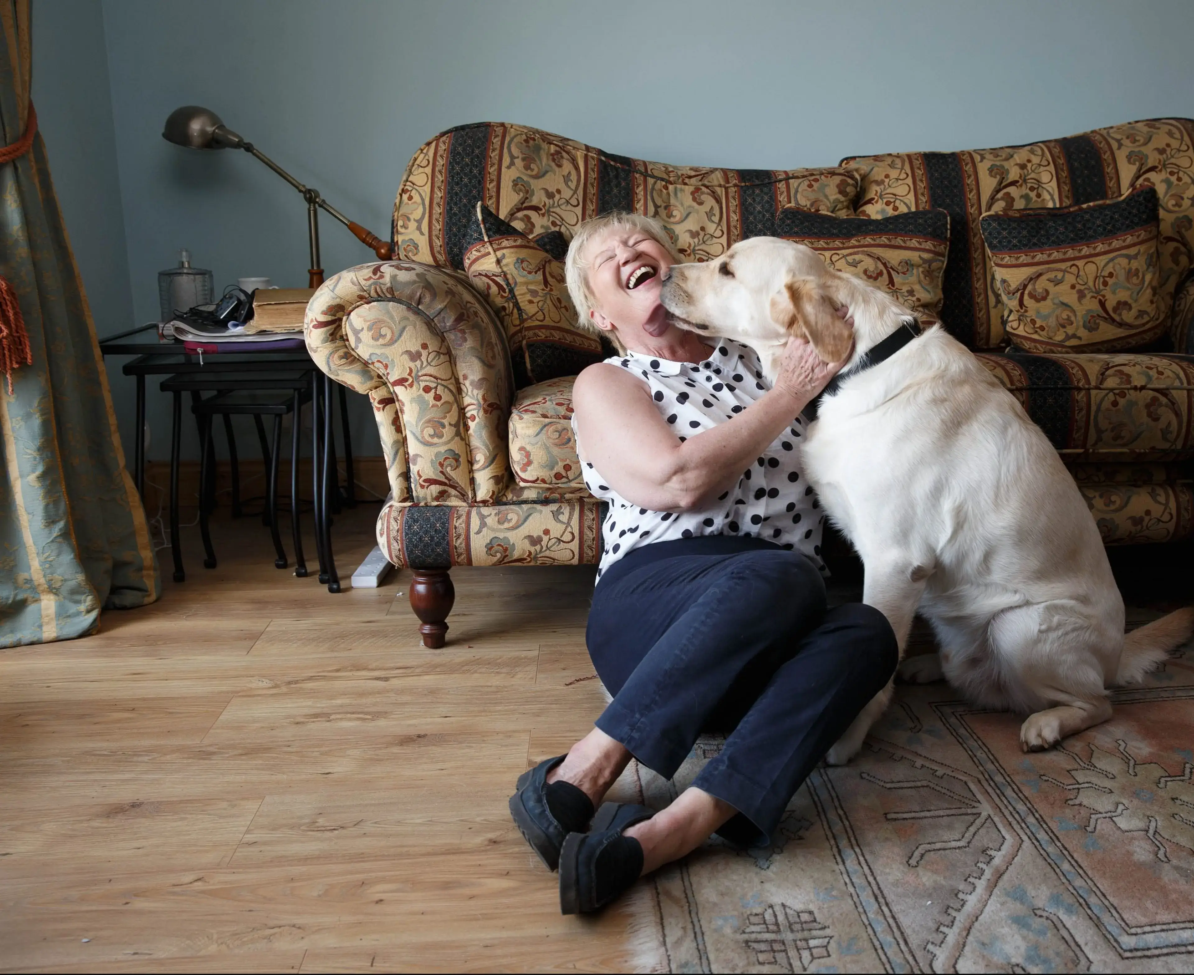 An elderly woman with white hair laughs joyfully as a white dog licks her face while she sits on the floor in front of an ornate patterned armchair in a living room.