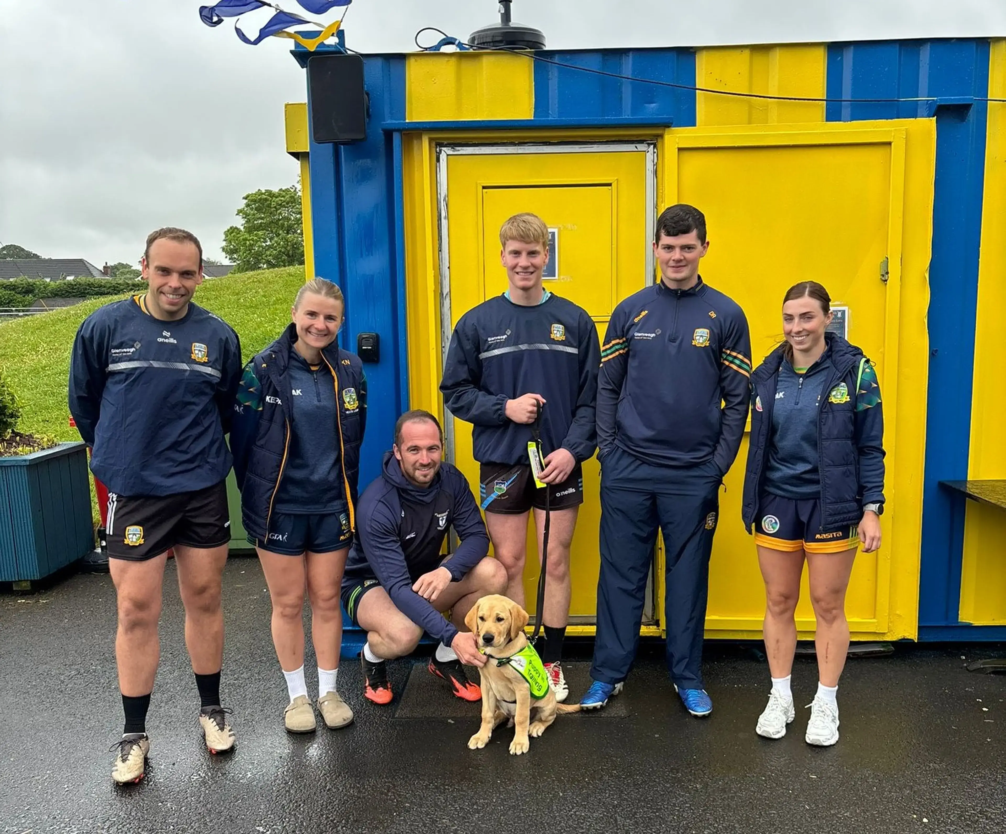 A group of six people in navy and yellow sports uniforms pose together in front of a blue and yellow container building, with a man crouching in the center holding a yellow Labrado