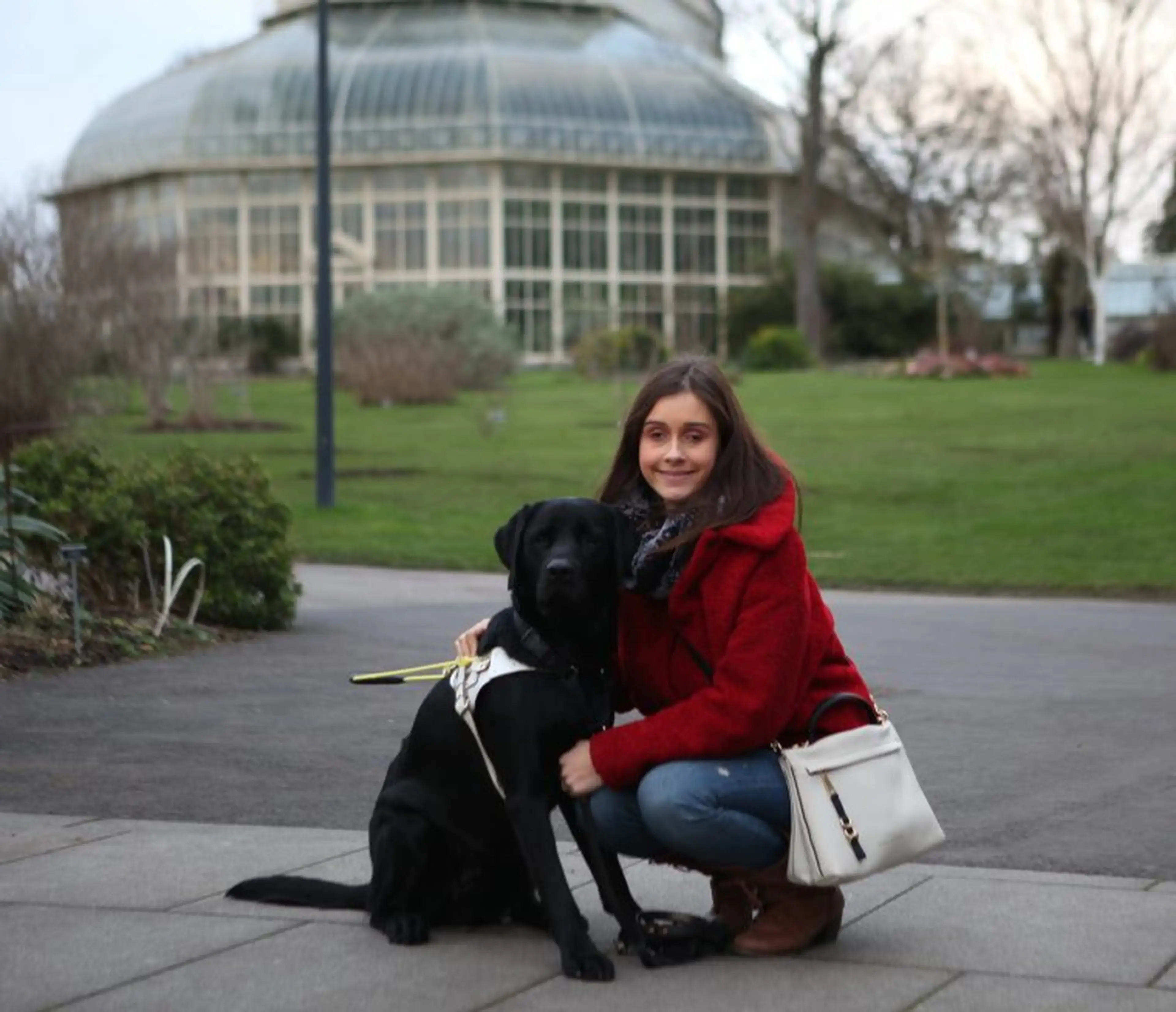 A young woman in a red jacket crouches beside a black Labrador dog on a paved path, with a large glass greenhouse building and manicured gardens visible in the background.