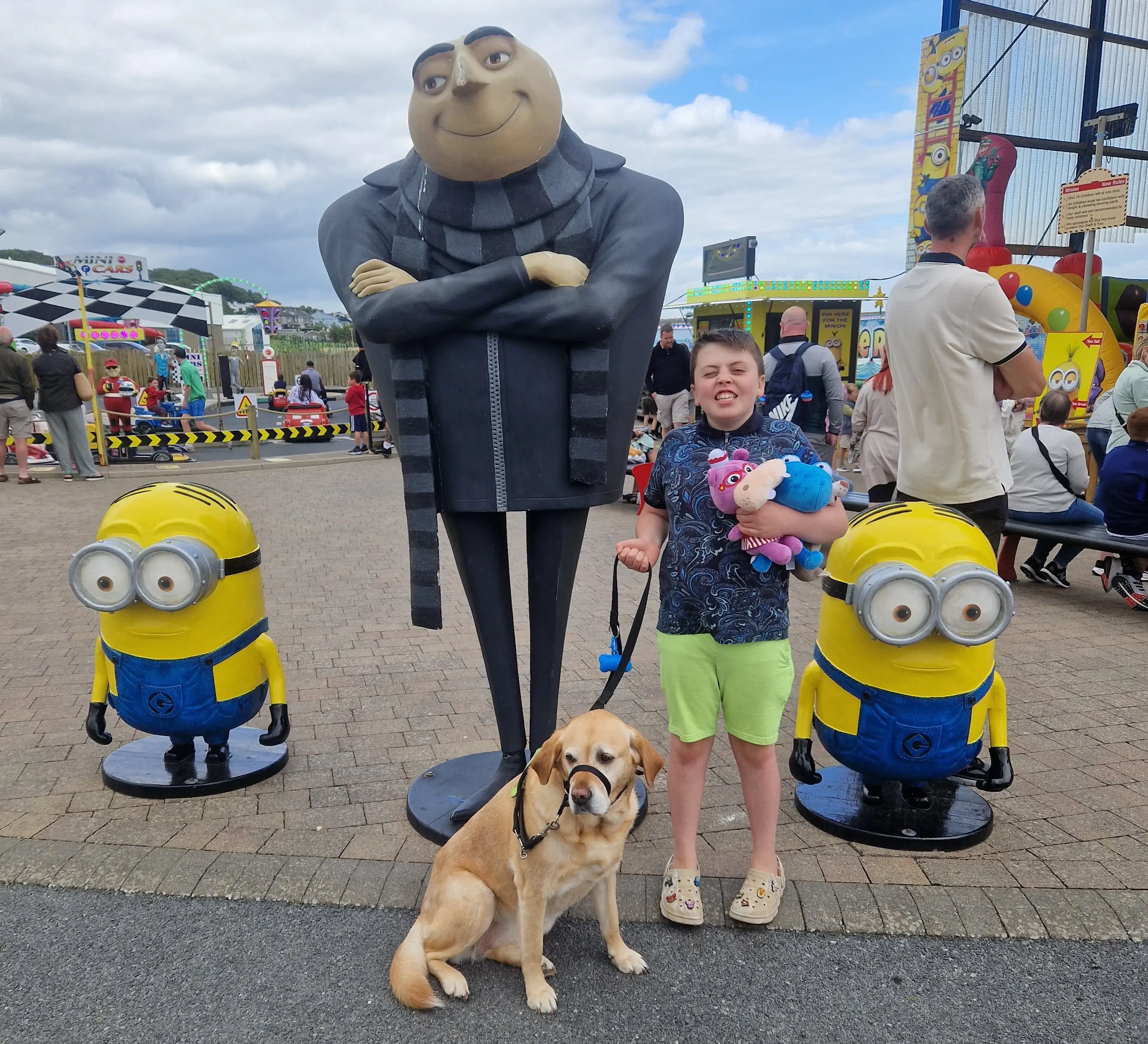 A young boy with a dog poses for a photo with life-sized character statues including Gru and two Minions from "Despicable Me" at an amusement park or carnival.