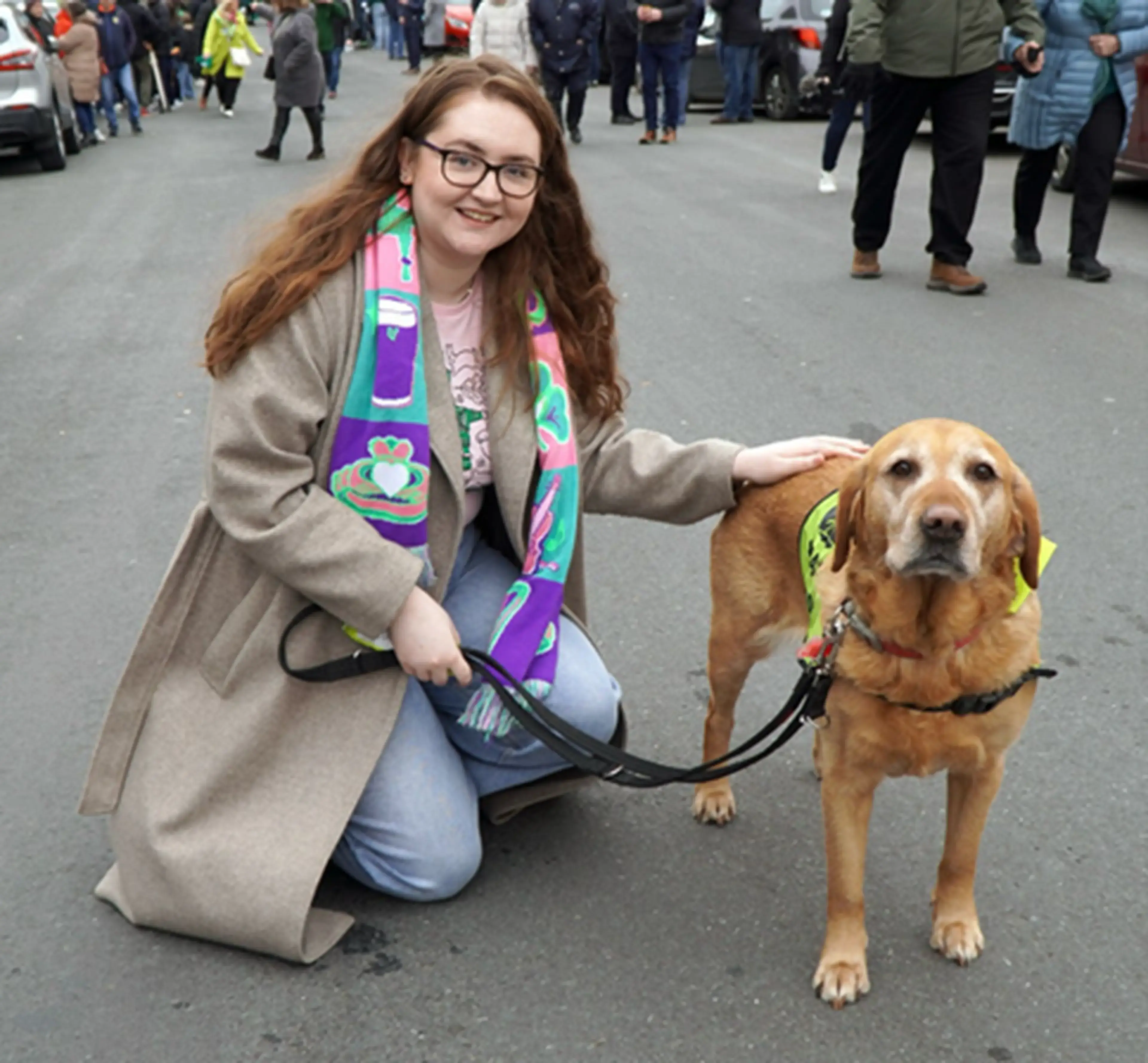 A young woman with long reddish-brown hair and glasses crouches beside a golden-brown Labrador Retriever wearing a yellow service vest on a crowded street.