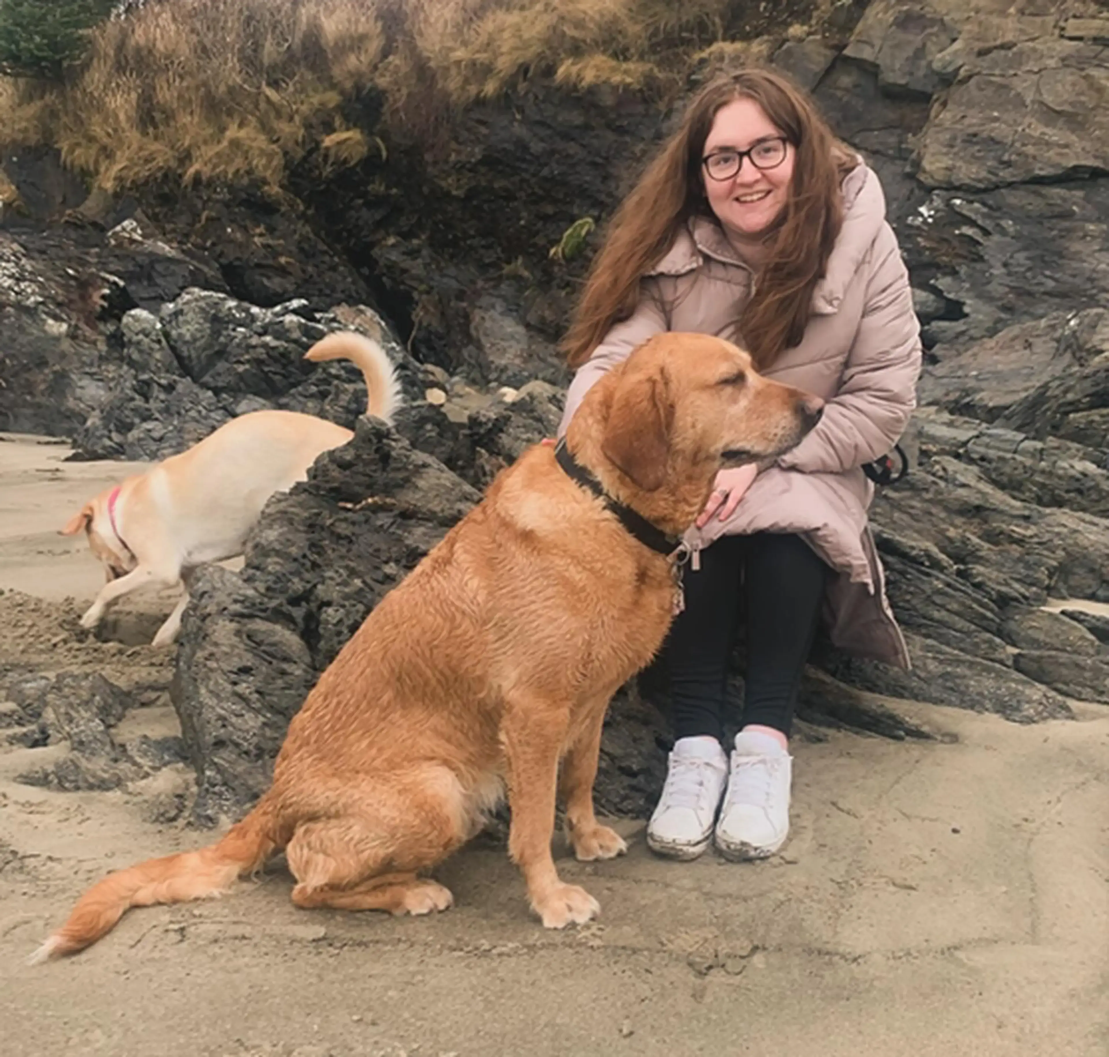 A smiling woman wearing glasses and a beige jacket crouches beside a brown dog on a rocky beach, with another light-colored dog visible in the background.