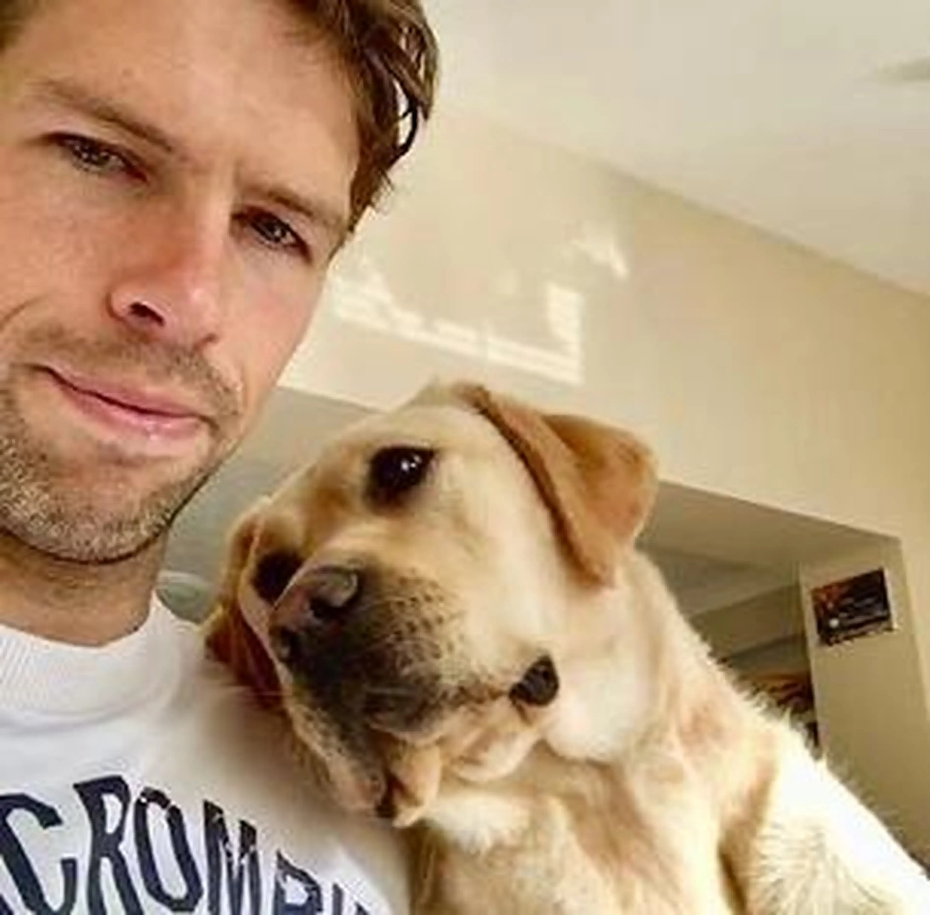 A man in a white t-shirt takes a selfie with a cream-colored Labrador Retriever indoors.