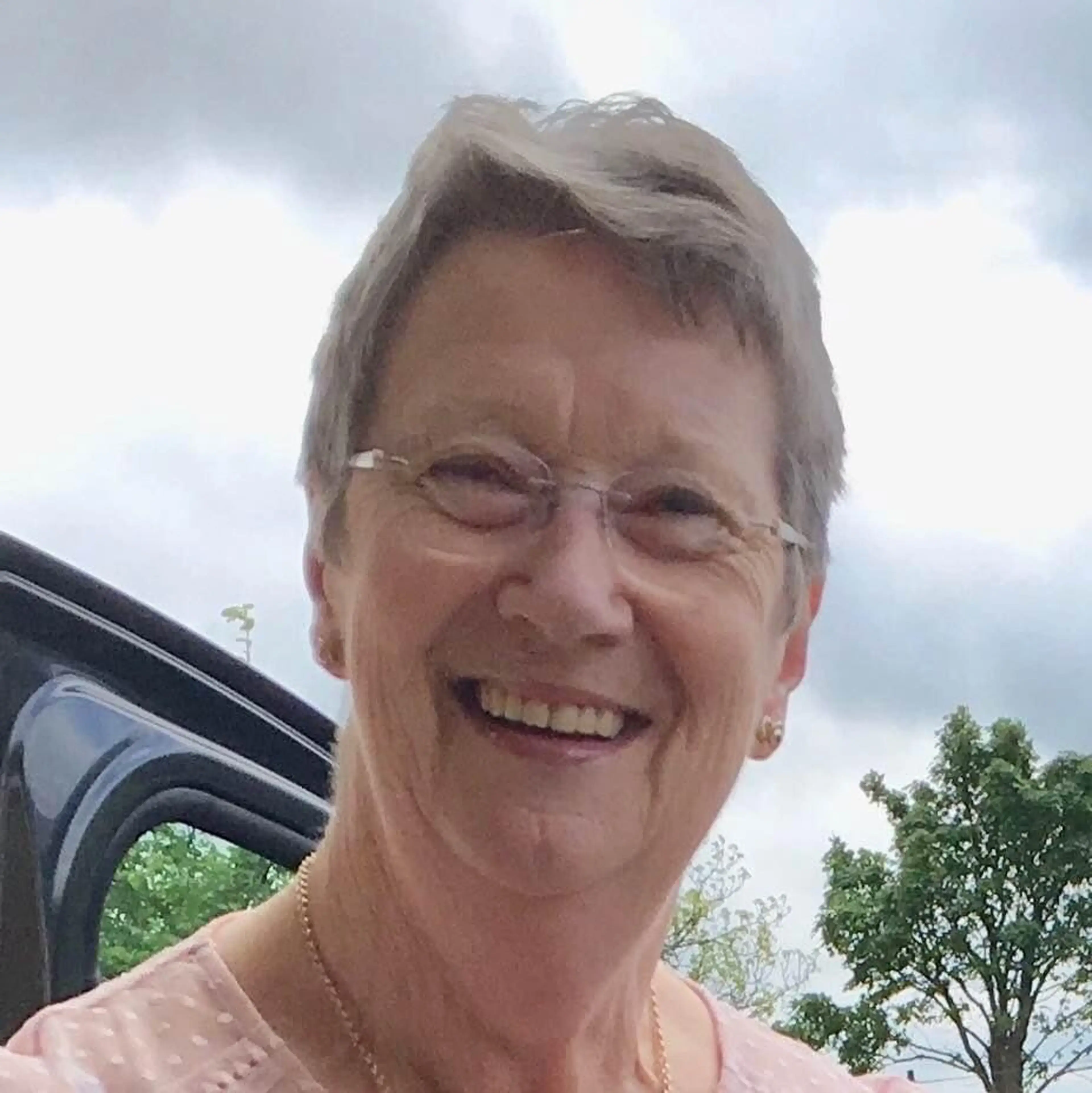A smiling woman wearing glasses and a pink shirt stands outdoors against a cloudy sky with trees visible in the background.