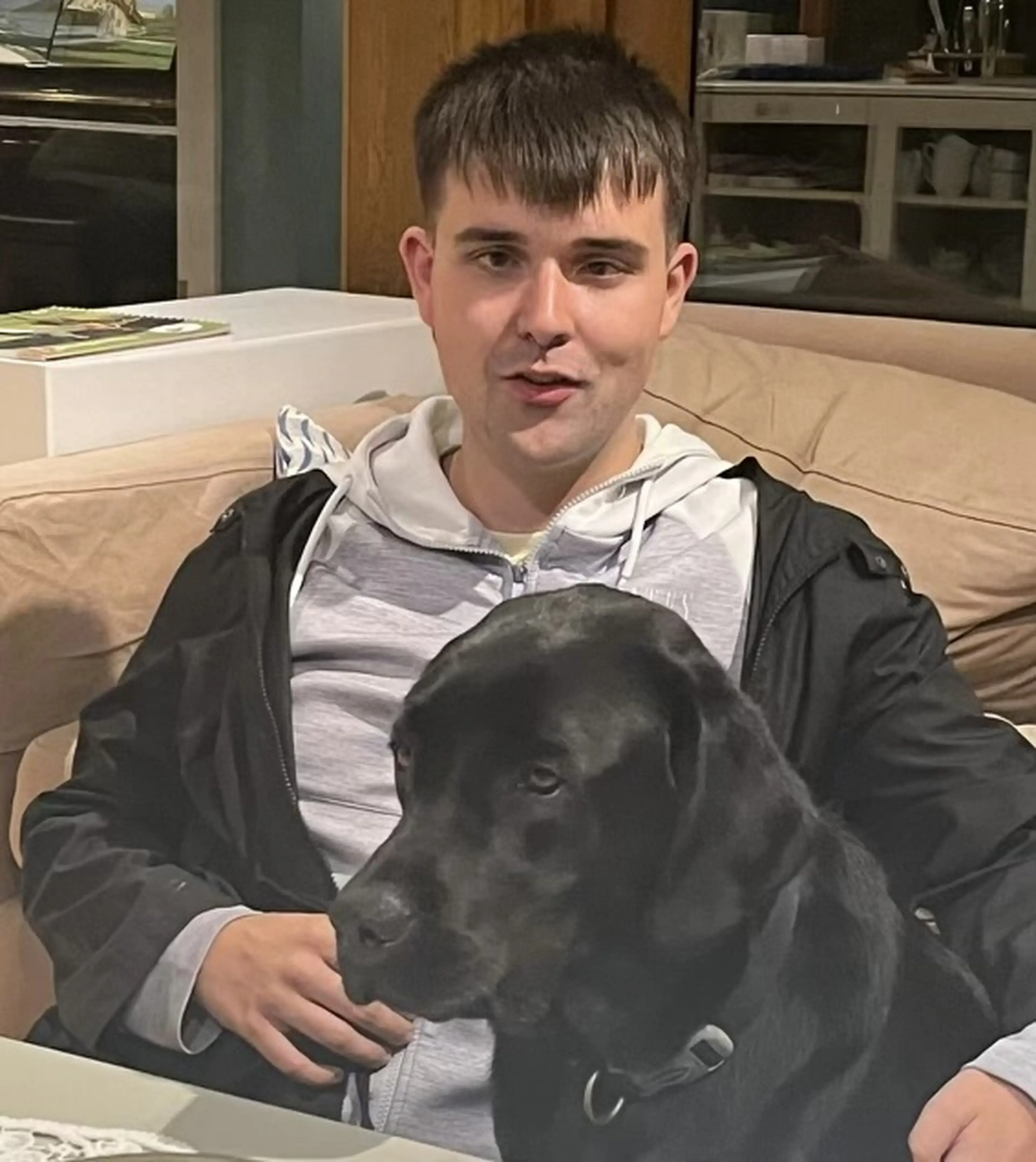 A young man sitting on a beige couch holds a black dog while speaking, with kitchen cabinets visible in the background.