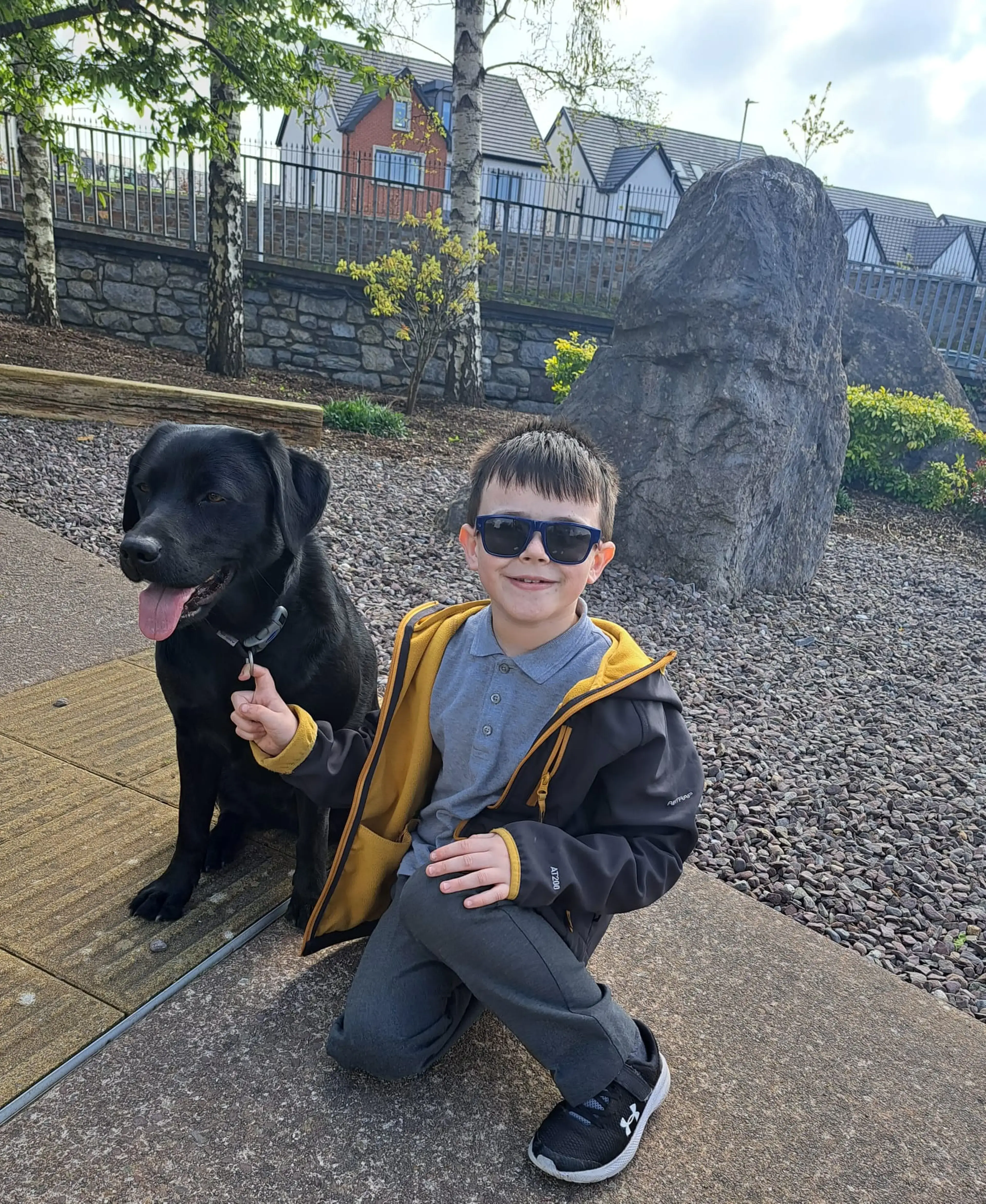 A young boy wearing sunglasses and a yellow and black jacket crouches next to a black Labrador dog on a path, with residential buildings and a large stone visible in the background