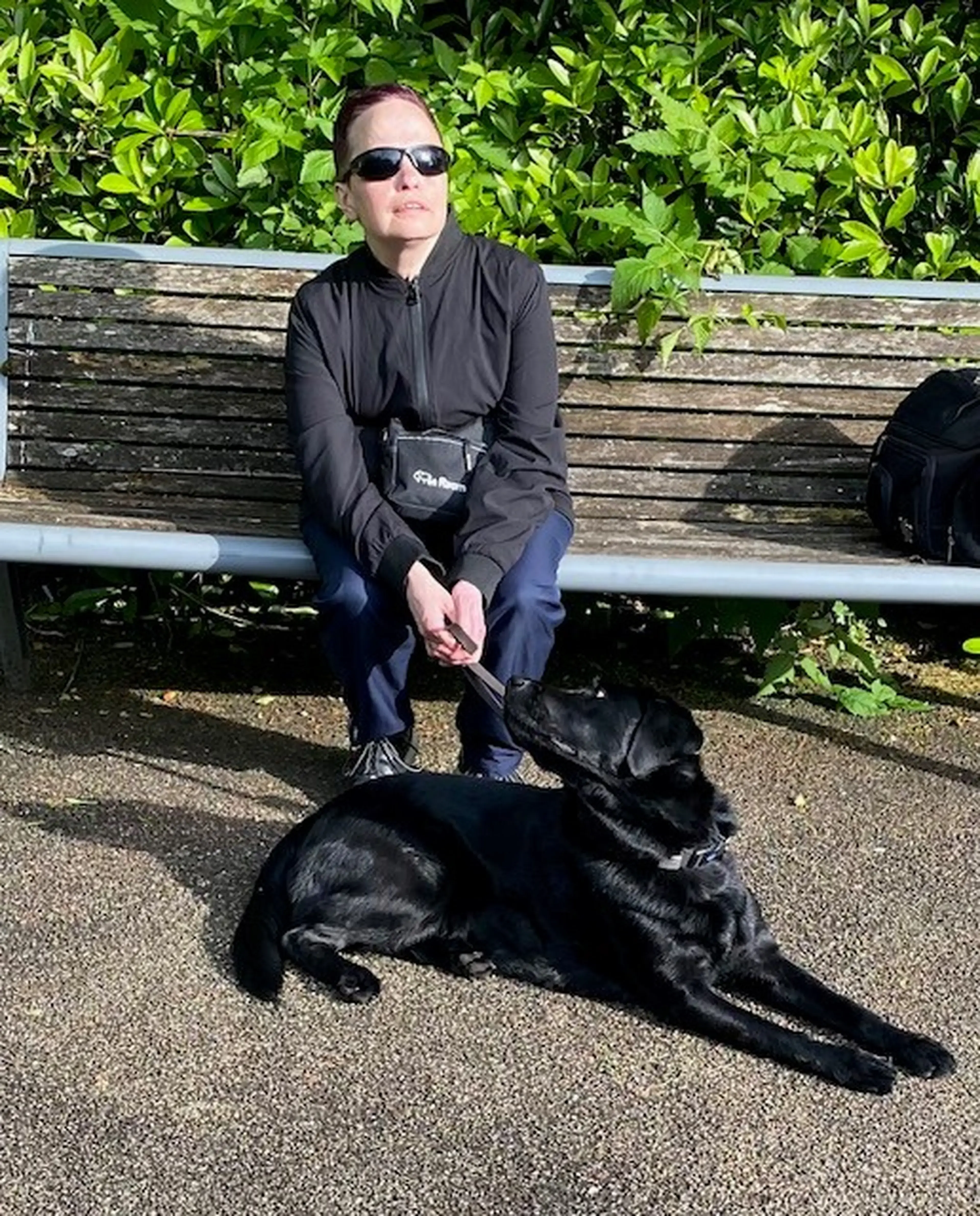 A person wearing sunglasses and dark clothing sits on a wooden bench while a black dog lies on the ground in front of them.