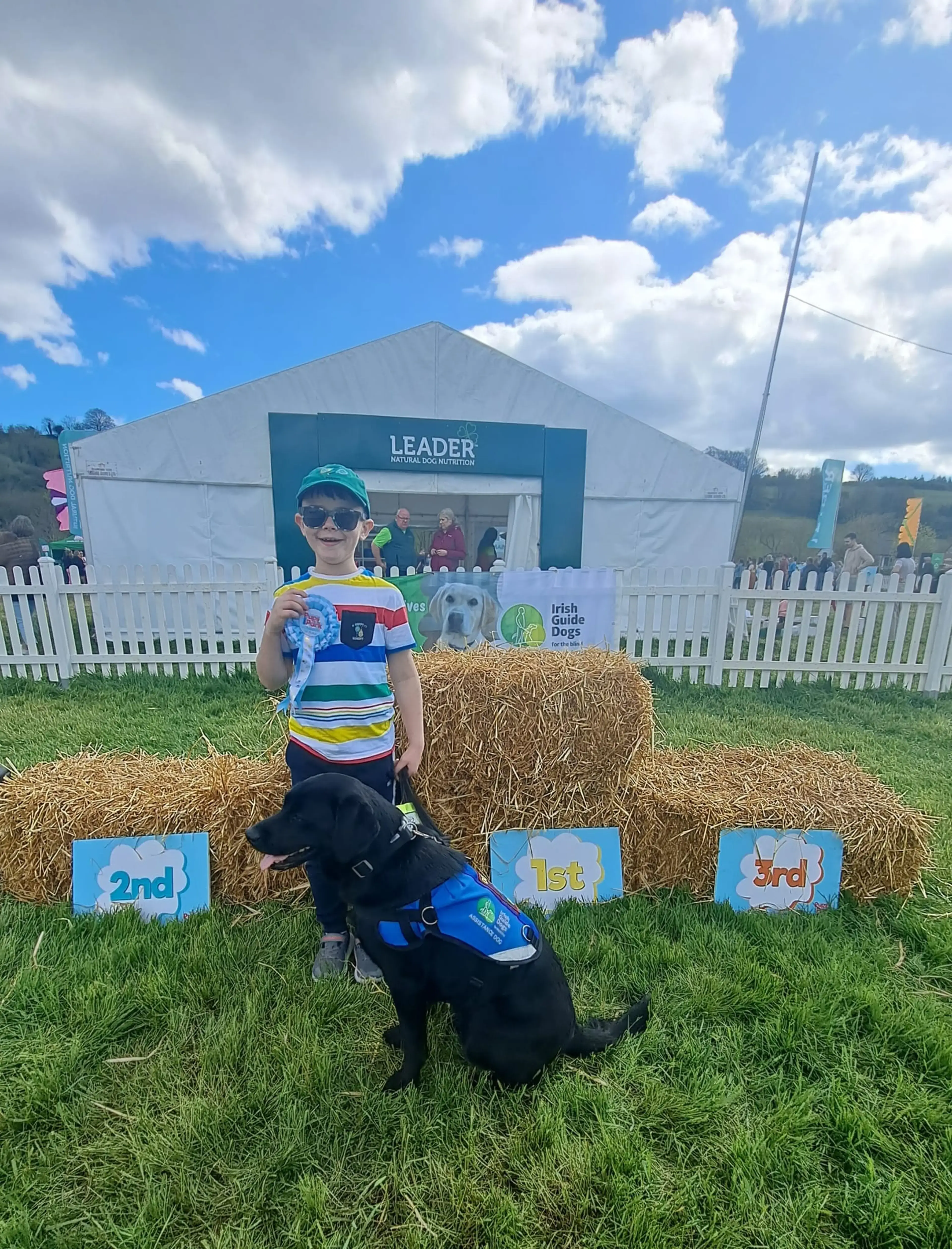 A young child wearing sunglasses and a colorful striped shirt stands with a black guide dog in front of a podium display at an outdoor event, holding a toy while posing for a photo