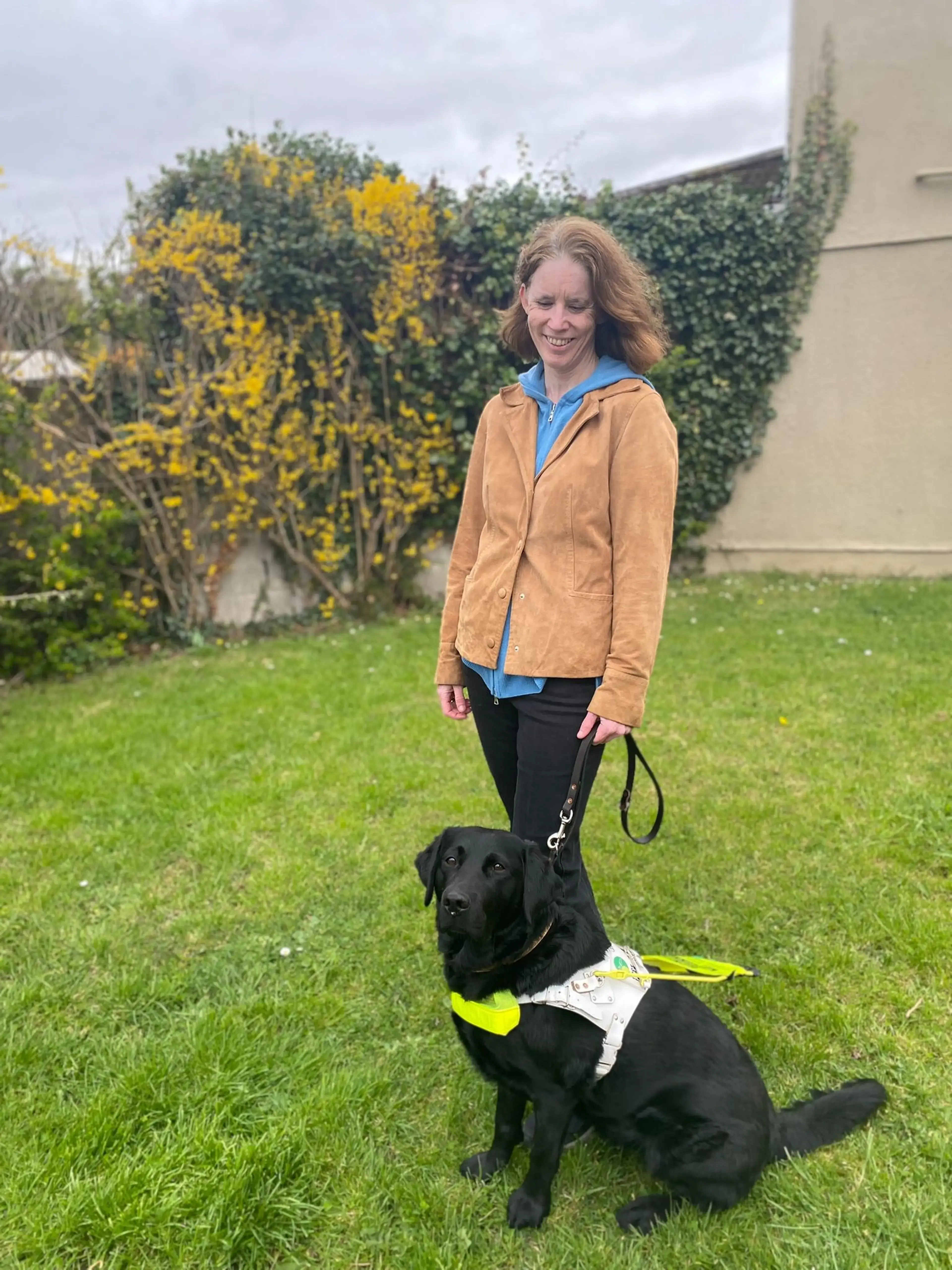 A woman in a tan jacket and blue shirt stands smiling in a garden while holding the leash of a black Labrador Retriever wearing a yellow and white service vest.