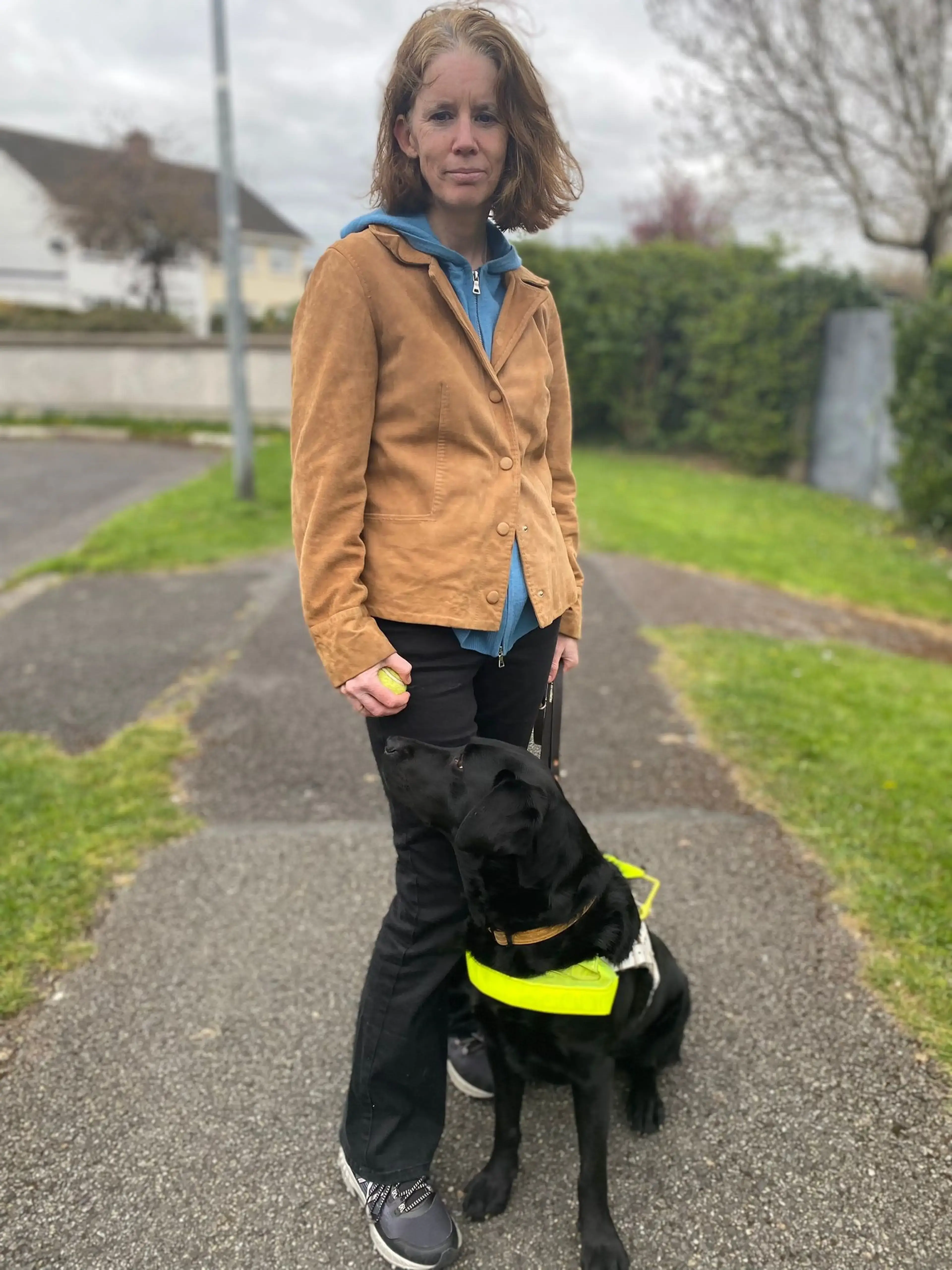 A woman stands on a residential path with a black Labrador Retriever wearing a high-visibility yellow vest, both facing the camera.