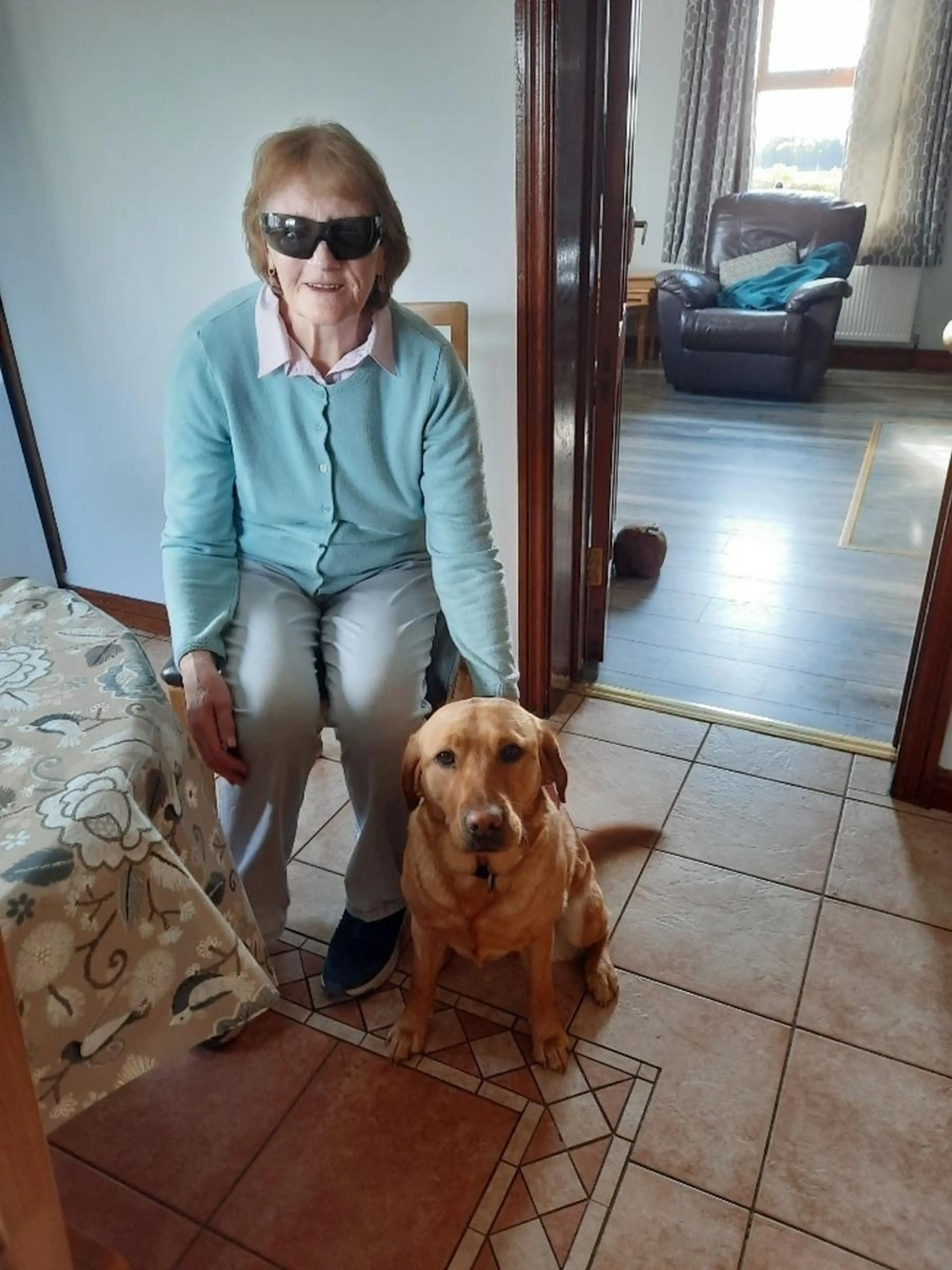 An older woman wearing sunglasses and a light blue cardigan poses indoors with a brown Labrador Retriever on a tiled floor.