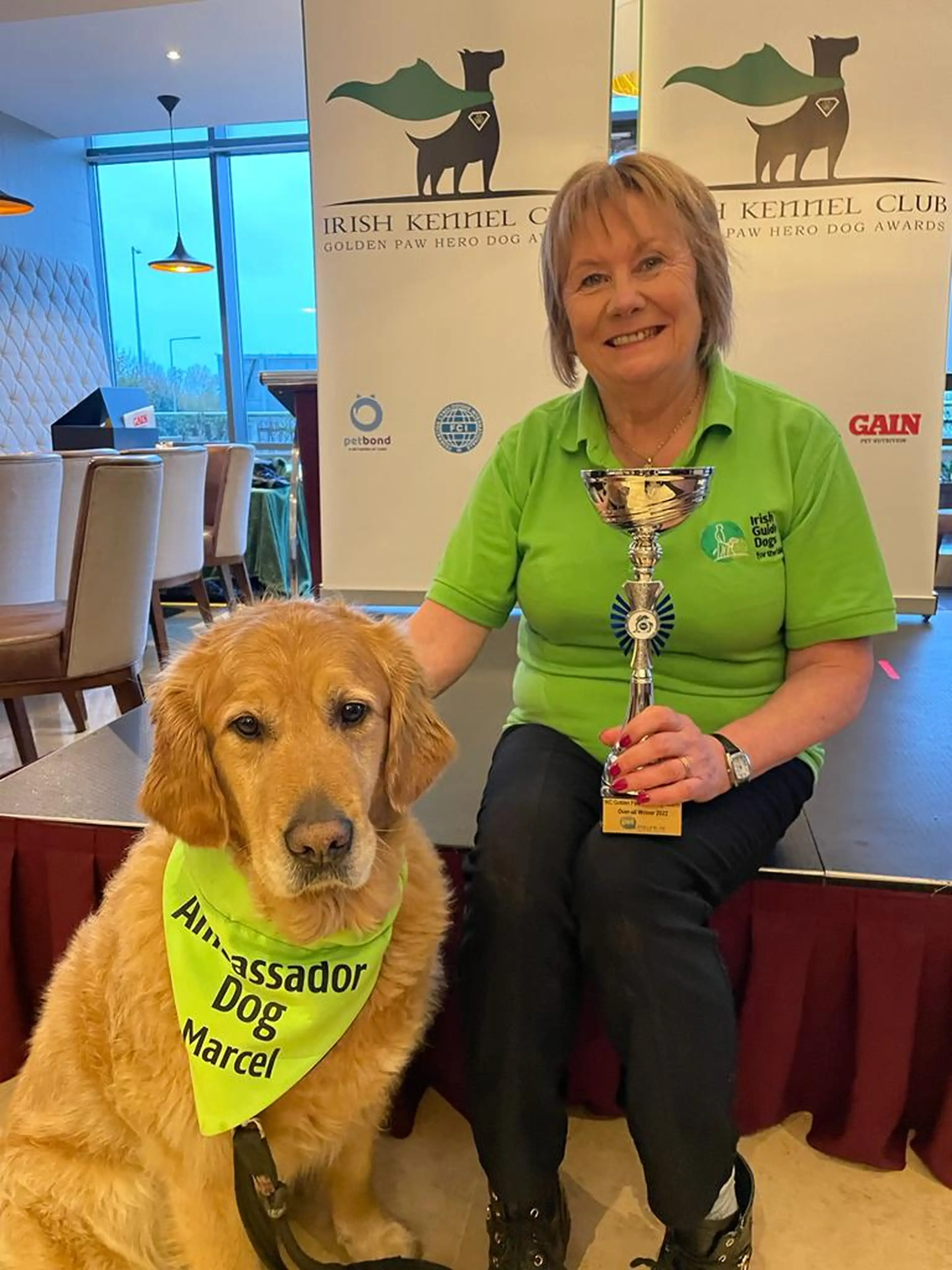 A woman in a bright green polo shirt holds a golden trophy while posing next to a golden retriever wearing an "Ambassador Dog Marcel" vest at an Irish Kennel Club Golden Paw Hero D
